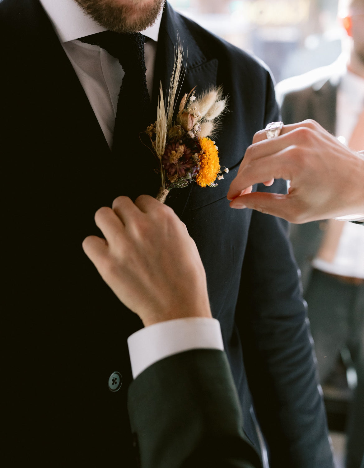 Person adjusting a boutonniere on a suit jacket. Dried flower buttonhole with yellow focal flower and dried grasses