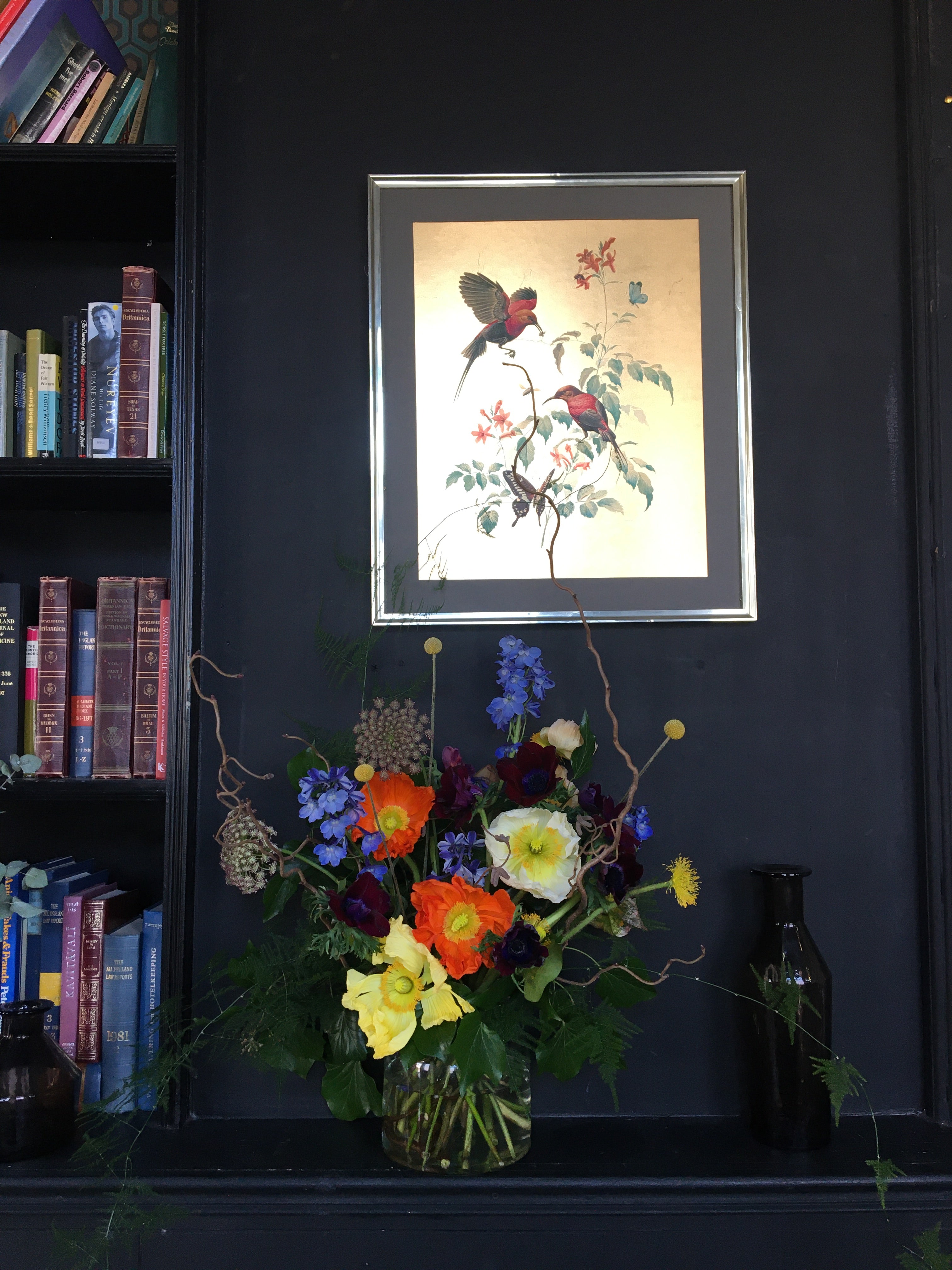 Framed artwork on a dark surface with a bouquet of flowers and books below. Colourful flowers in a vase of blue delphinium, burgundy anemone, cream and pale yellow poppies contrast against the dark wall