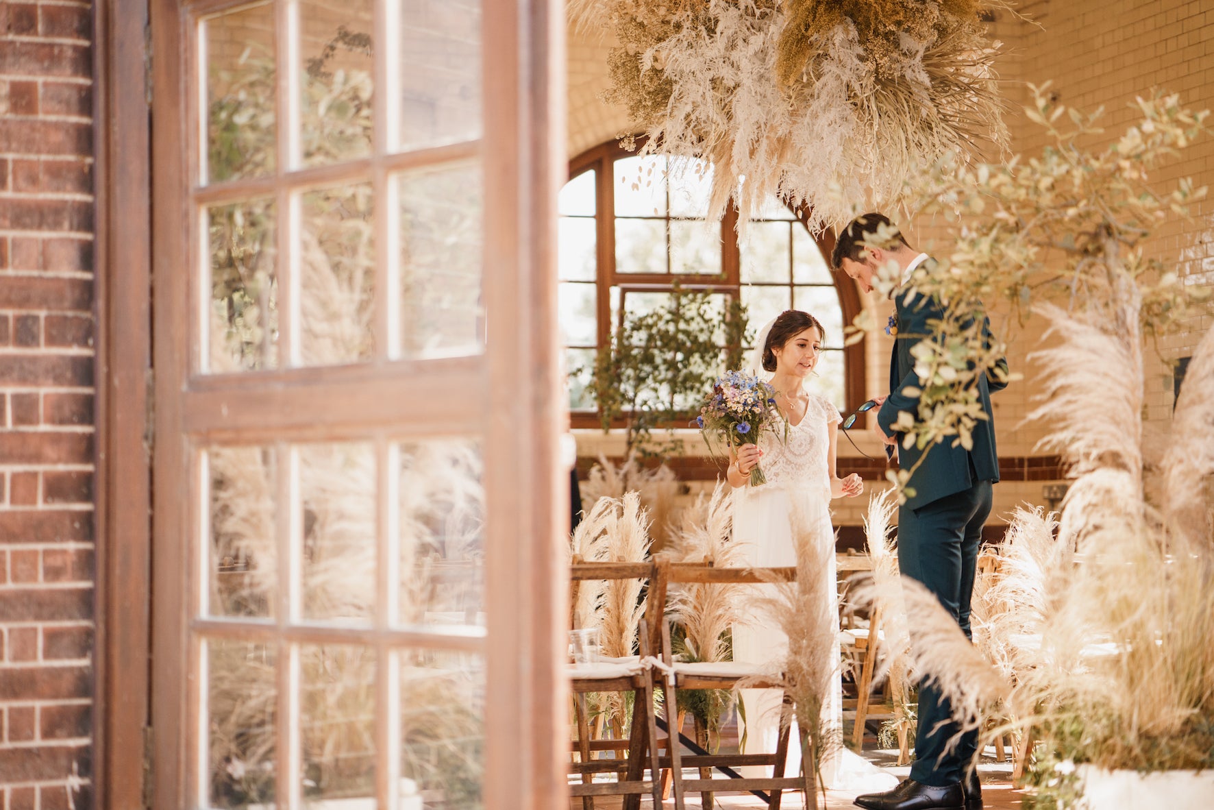 Wedding couple standing in a decorated indoor setting with pampas grass and large windows.