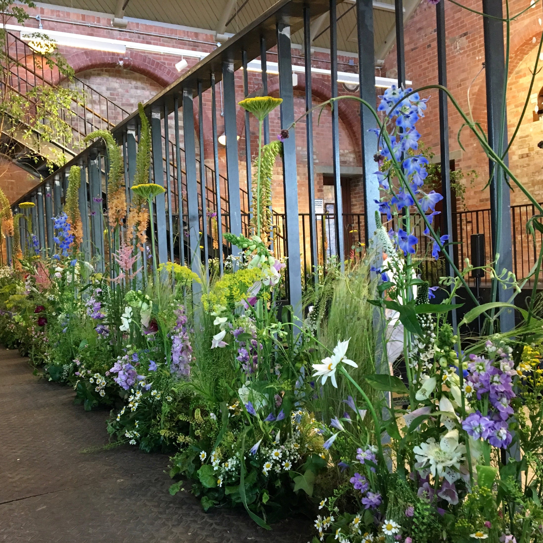 A lush garden bed filled with tall green foliage, purple delphiniums, white lilies, and yellow flowers lining a walkway under a brick arched gallery structure.