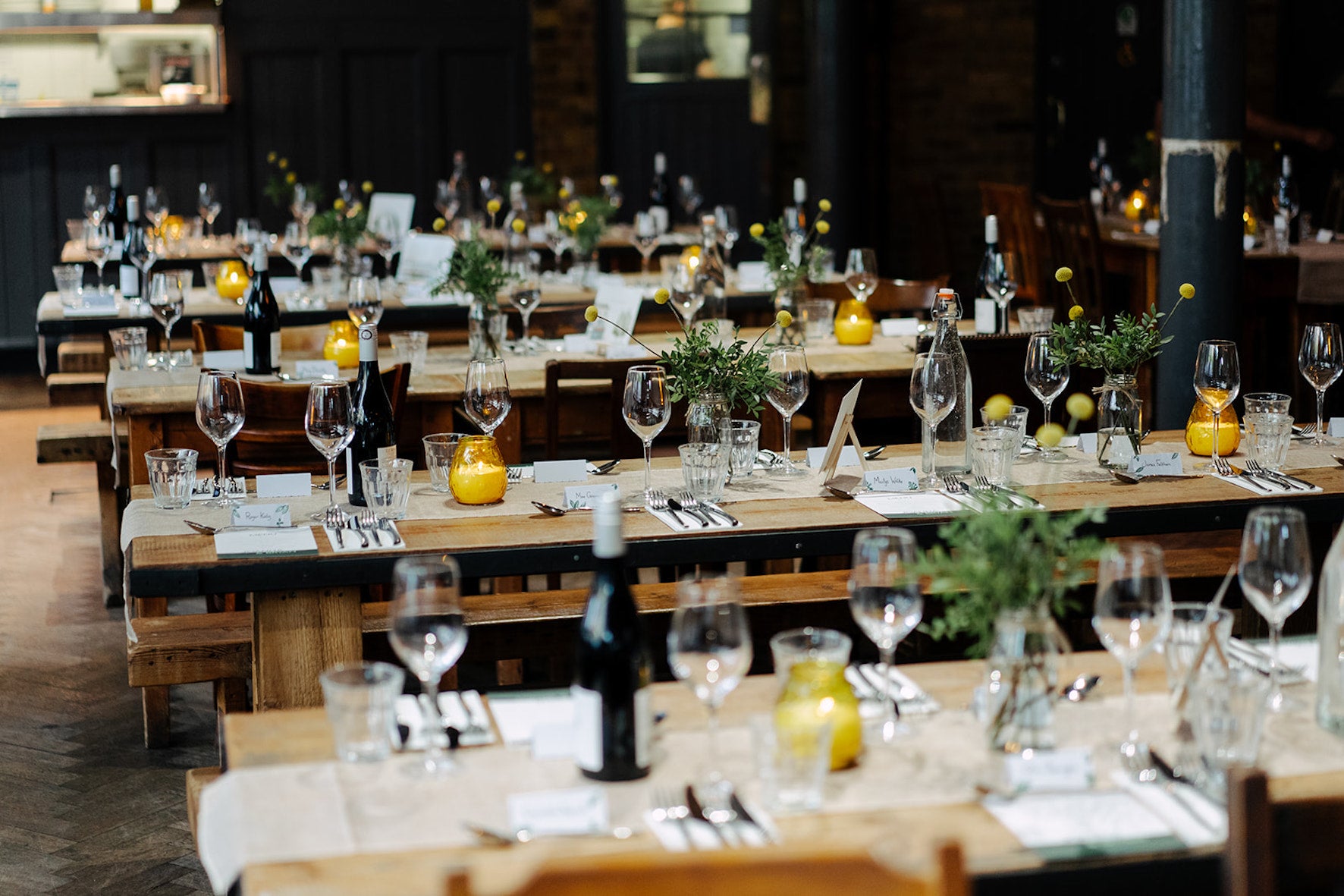 Decorated tables with wine glasses, candles, and greenery and yellow flowers in a banquet hall.