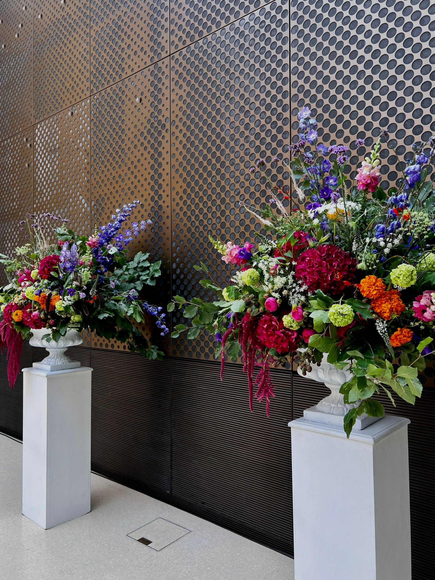 Colourful floral arrangements on white pedestals against a patterned wall. Bright summer flowers including hydrangea, Guelder rose, Delphinium, carnation, roses and Ammi