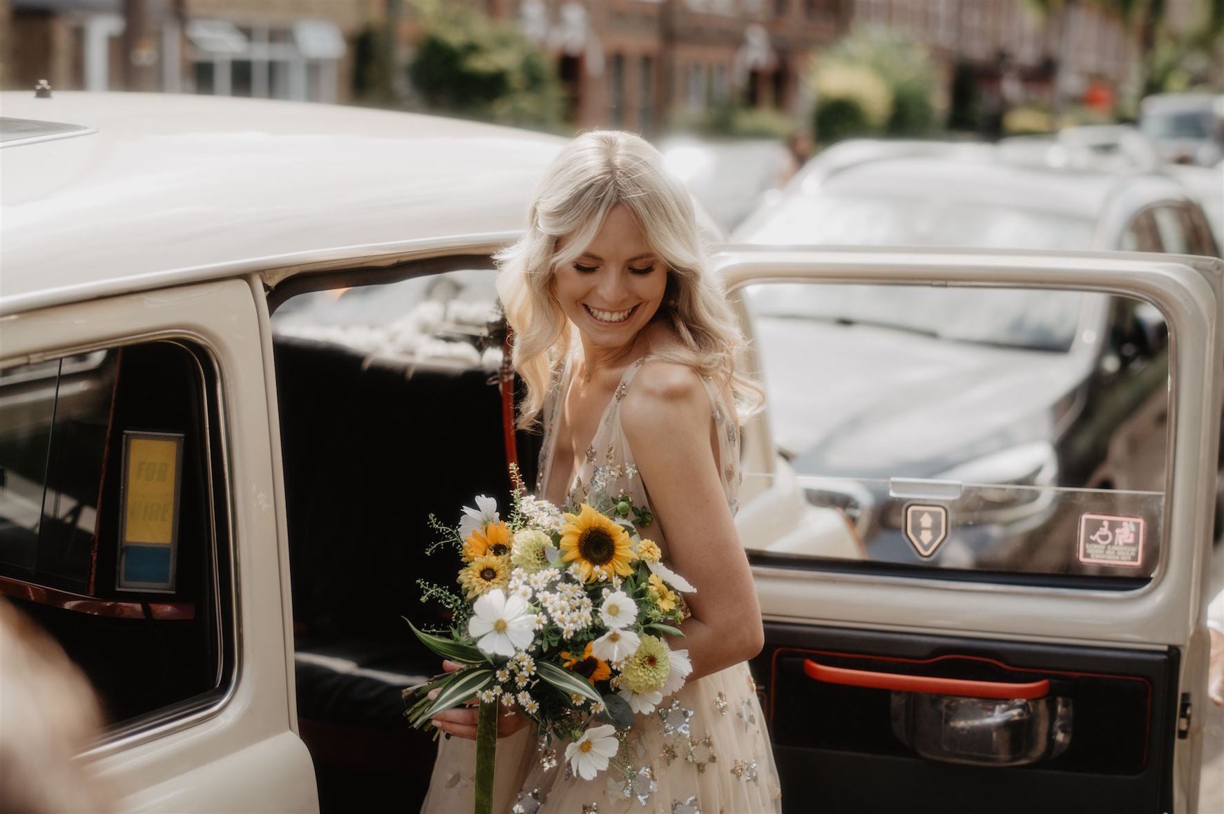 Bride in a wedding dress holding a bouquet of flowers, standing next to a vintage taxi. Bridal bouquet of sunflowers, white cosmos, daisies, zinnia and grasses