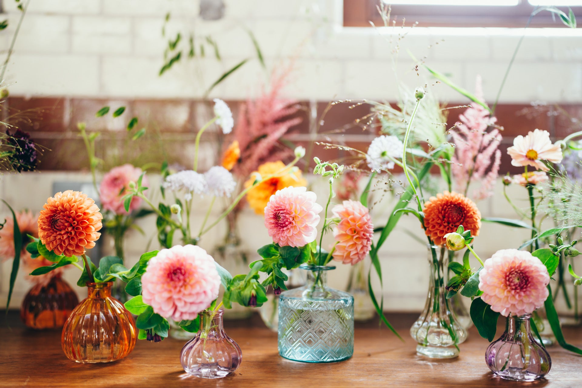 Decorative arrangement of flowers in bud vases on a wooden surface with a blurred background. Flowers are dahlia in pastel pink and orange with grasses