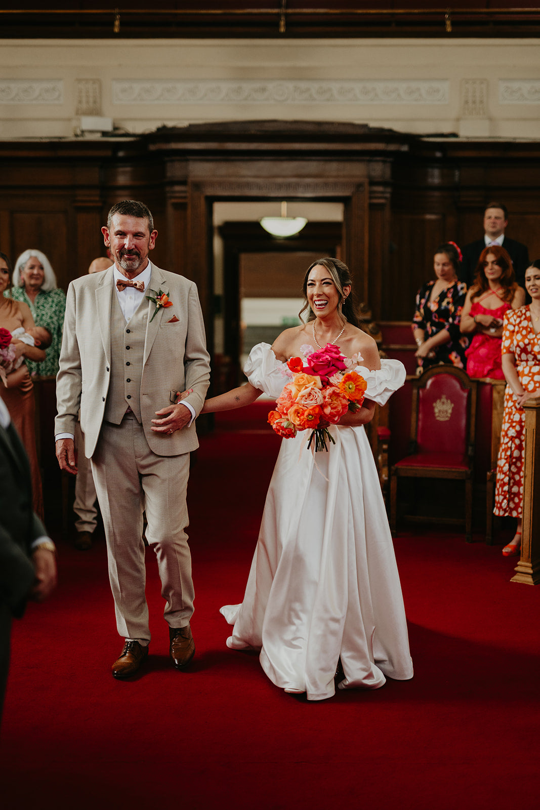 Wedding couple walking down a red carpet with guests in formal attire. Brightly coloured bridal bouquet of coral peonies, pink roses and orange icelandic poppies