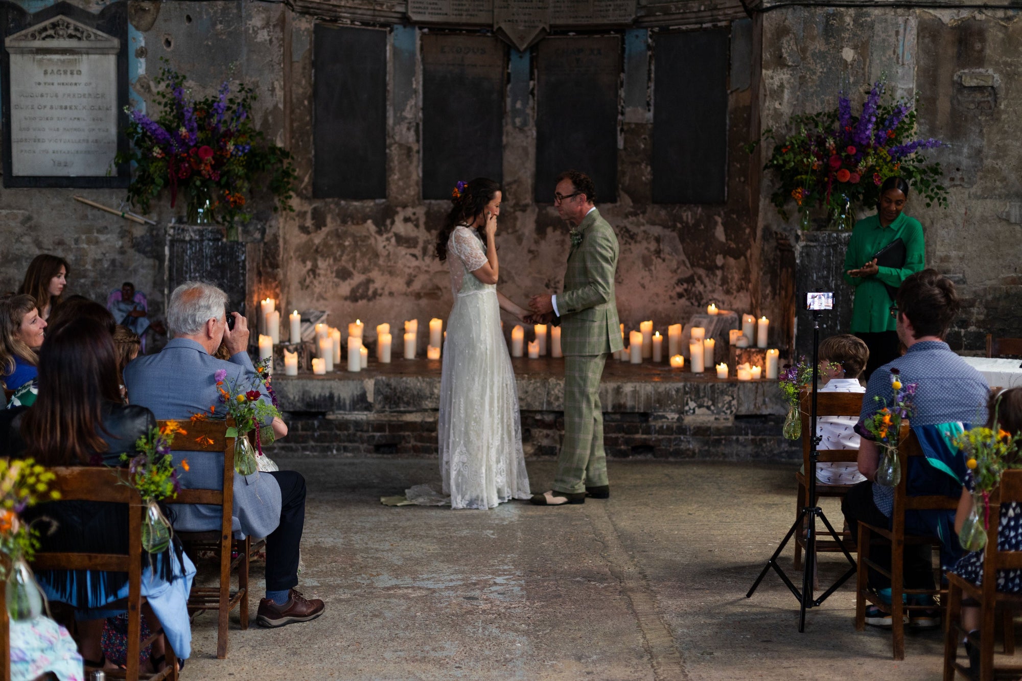 Wedding ceremony taking place in a rustic indoor setting with candles and floral arrangements. Large pedestal arrangements are brightly coloured in purple and pinks. Garden and meadow flowers are tied to the chairs along the aisle
