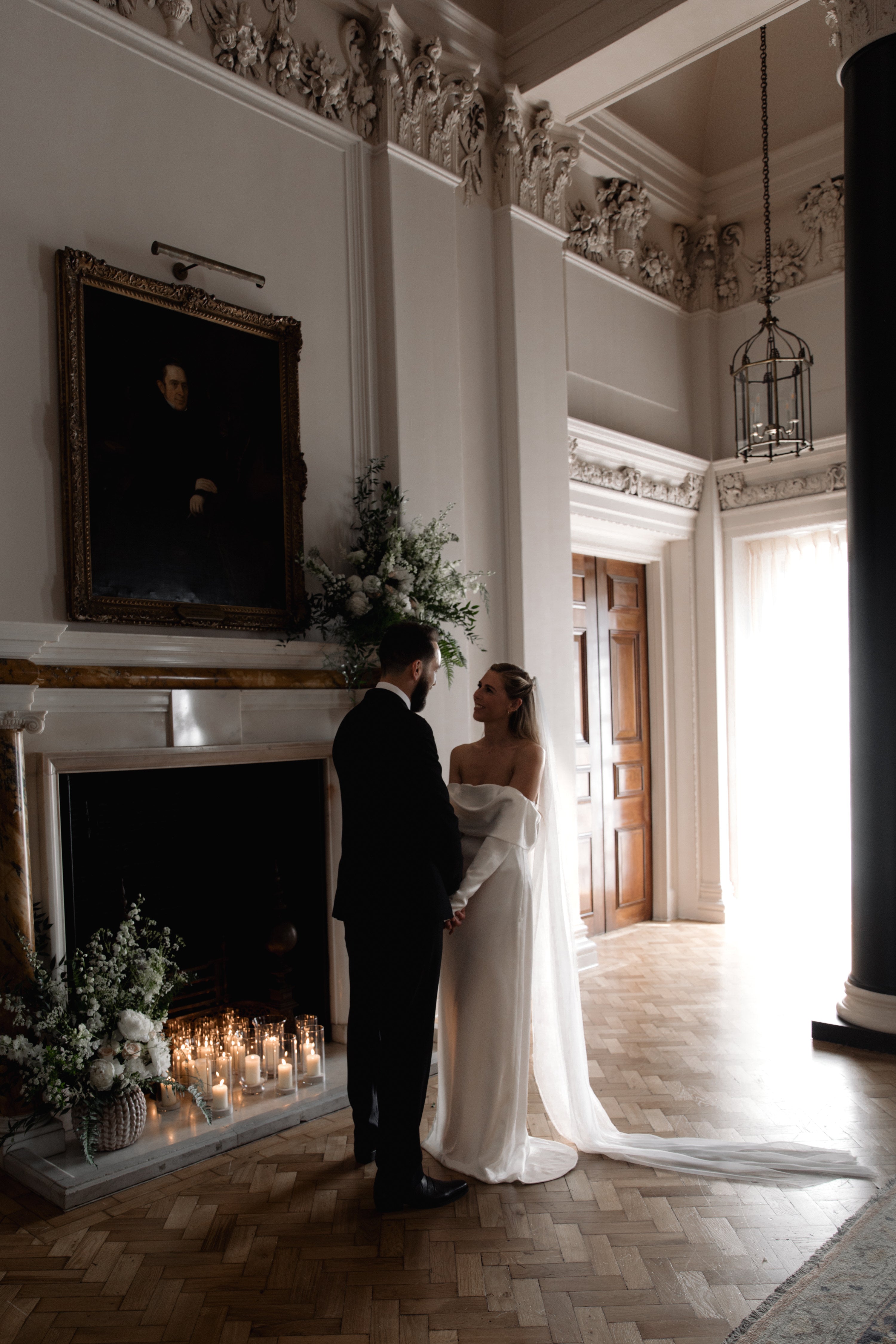 Wedding couple standing in a grand room with a fireplace and decorative elements. Romantic white and green floral arrangements decorate the fireplace and include delphinium, roses and peonies