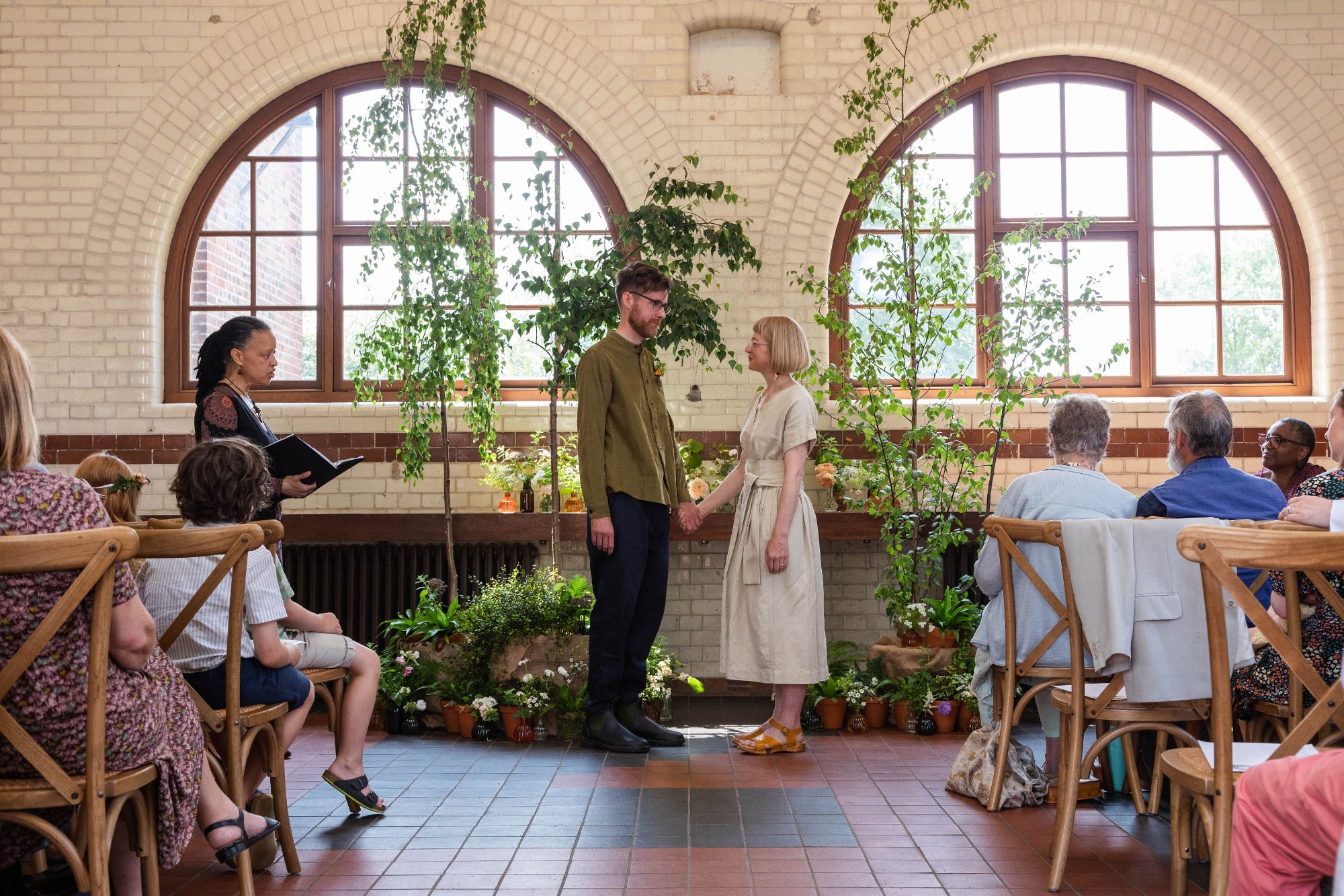 Bride and groom holding hands in a rustic room with seated guests. There is a backdrop of tall birch trees with potted plants and small vases of flowers at the base