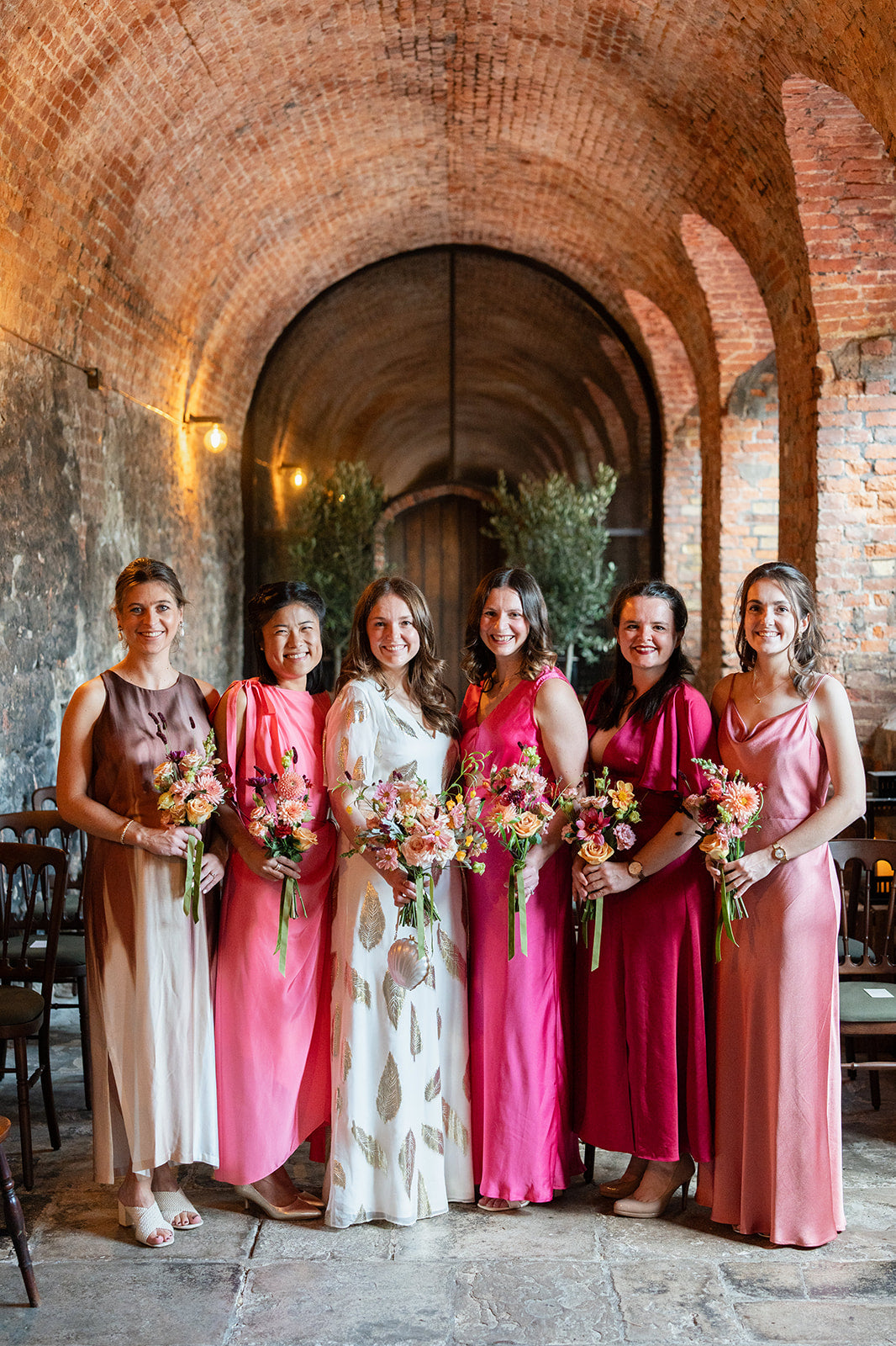 Bride and bridesmaids in colorful pink tone dresses standing in a brick archway.Each lady holds a bouquet of bright and pastel pink romantic stlye garden flowers