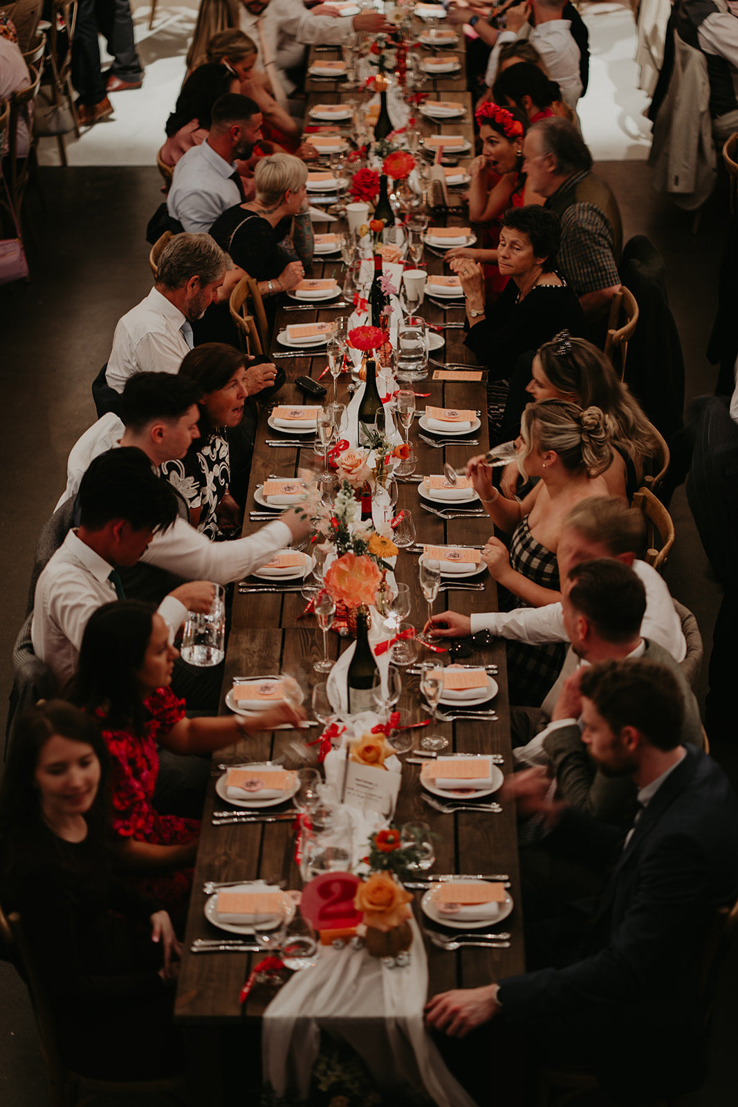 Wedding party dinner with guests seated around a long table set for a meal. Bright coloured peonies and roses add colour down the centre of the table