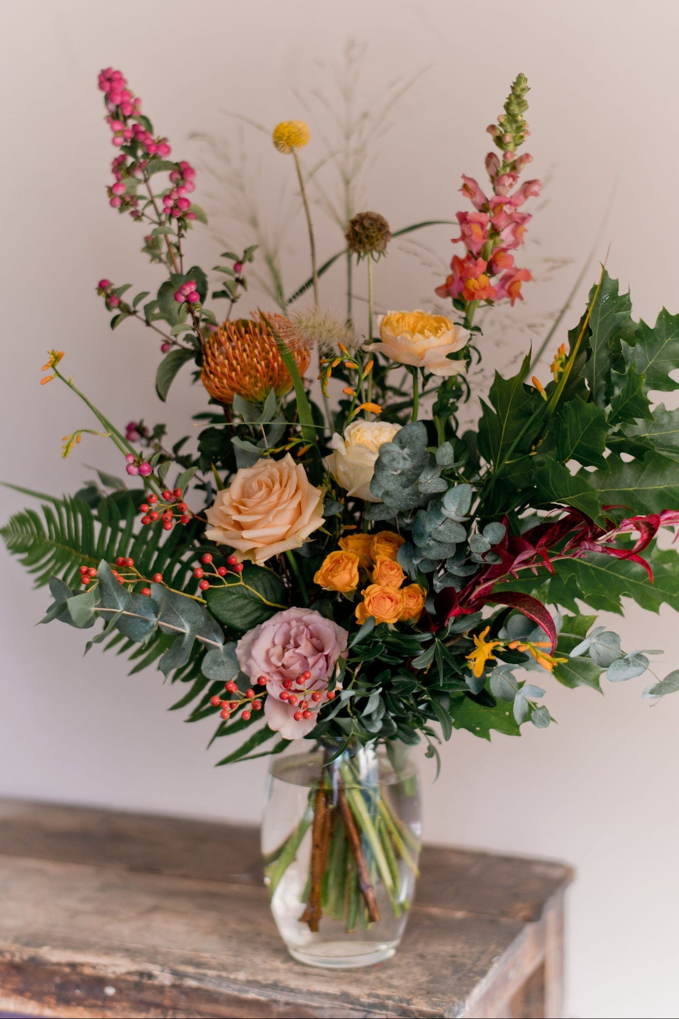 A vibrant, rustic-style floral arrangement in a clear glass jar vase, featuring a mix of orange, pink, yellow, and green flowers with textured foliage, displayed on a weathered wooden table against a neutral beige wall.