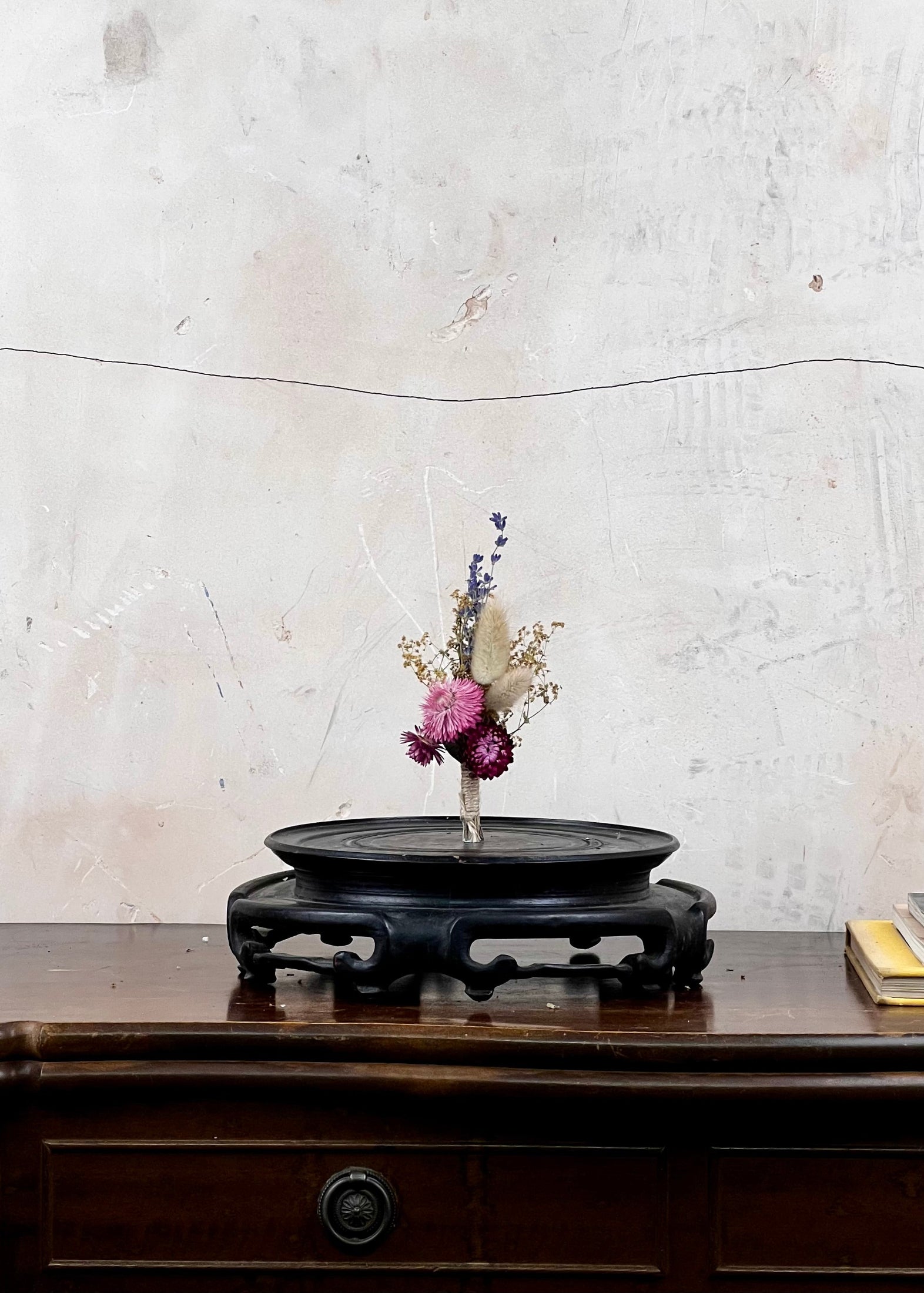 Dried flower buttonhole on a dark wooden stand against a textured white wall.