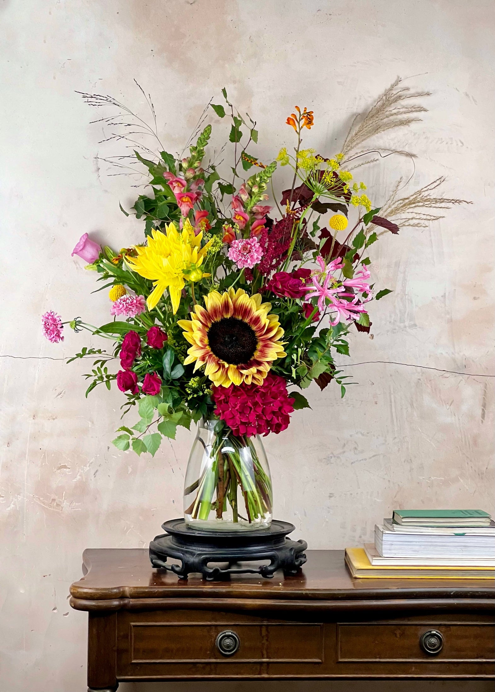 A vibrant, multicolor floral arrangement featuring a large yellow and brown sunflower, pink and red roses, yellow and pink chrysanthemums, and pampas grass, displayed in a clear glass vase on a black stand atop a dark wood console table.