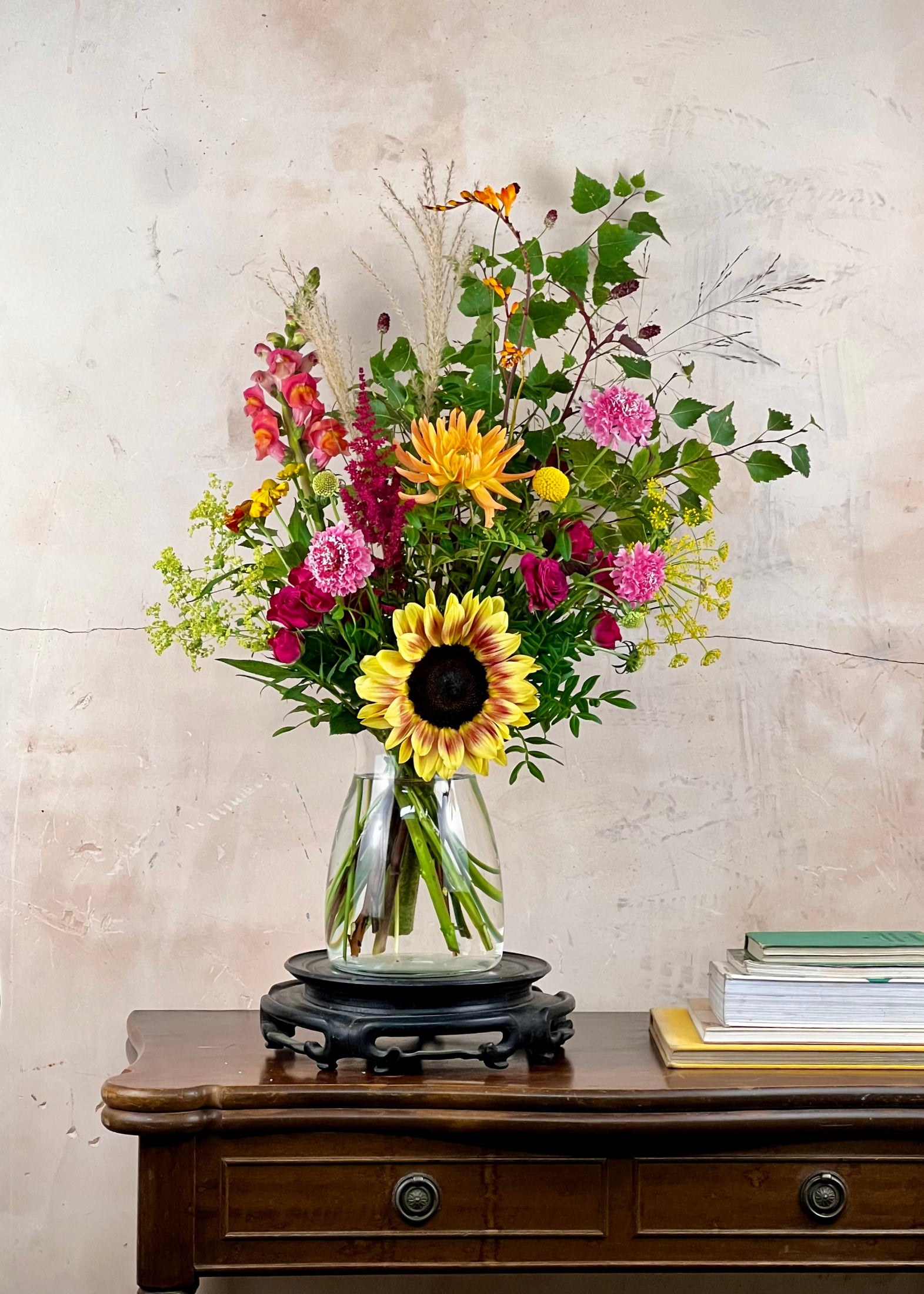 A vibrant mixed floral arrangement featuring a large yellow sunflower, pink and red flowers, and green foliage, displayed in a clear glass vase on a black wooden stand atop a dark wood console table against a textured beige wall.