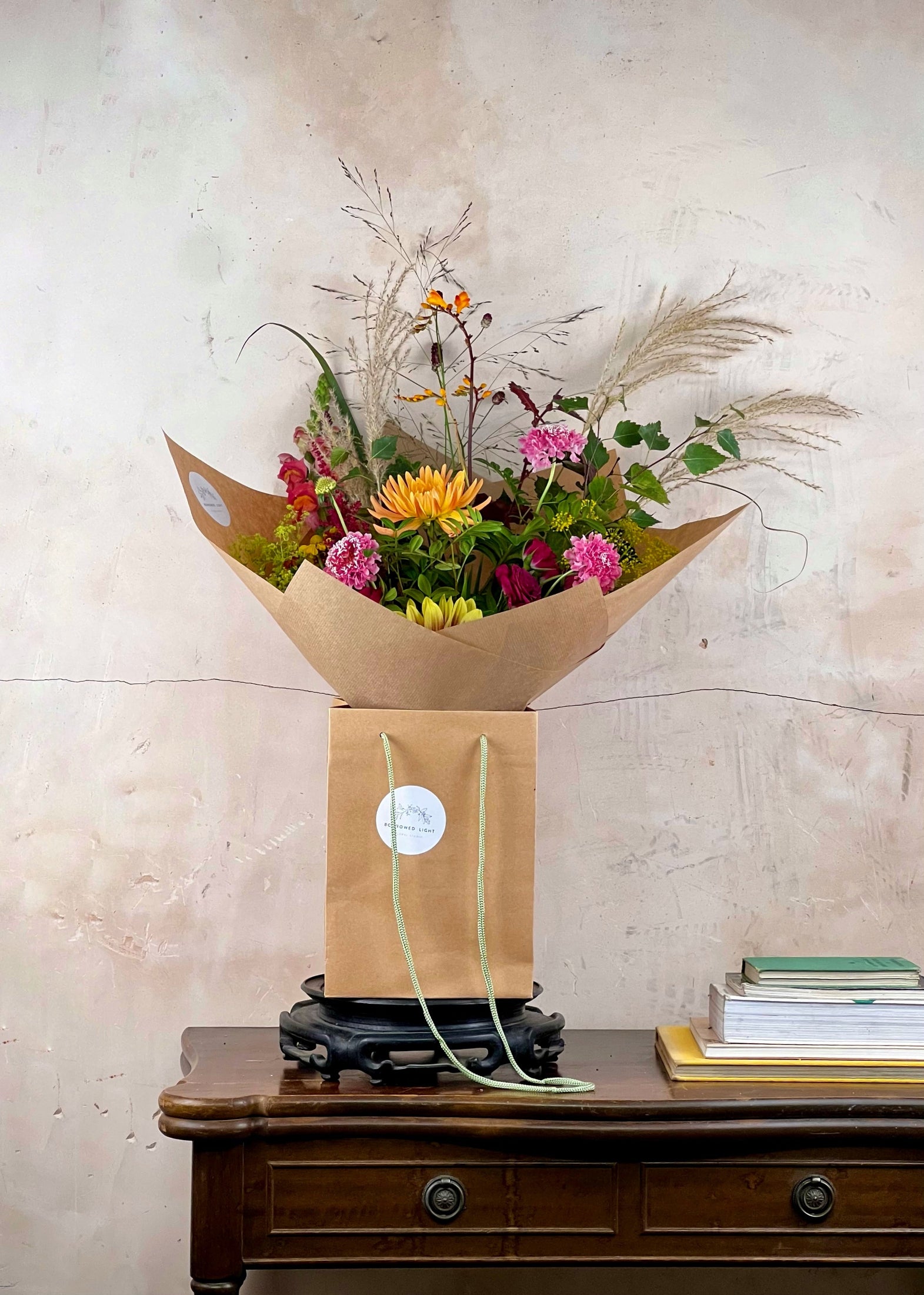 A floral arrangement in a kraft paper bag with paper handles, displayed on a dark wooden table against a textured beige wall. The bouquet includes orange, pink, and yellow flowers with dried pampas grass.