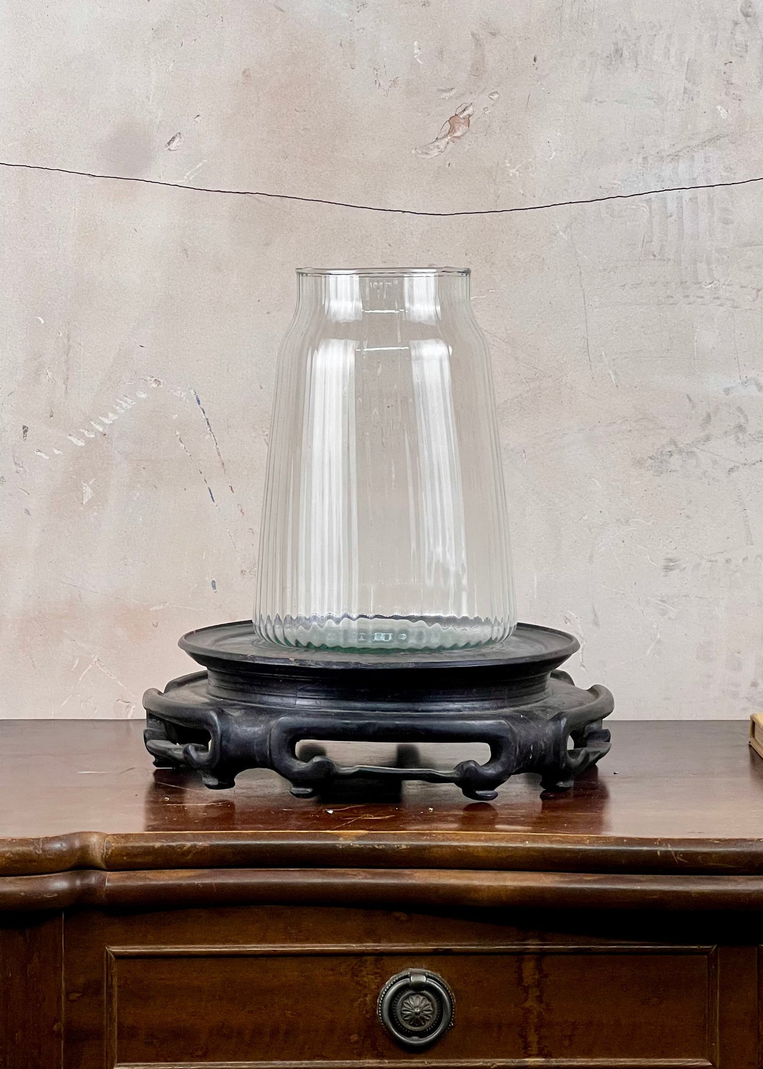 Clear recycled ribbed glass vase n on a wooden stand against a textured wall.