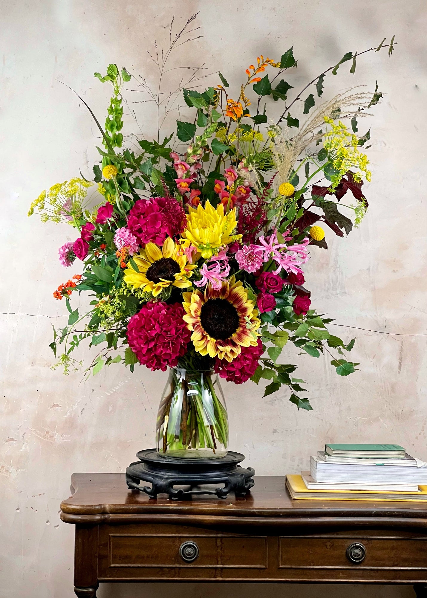 Colourful flower arrangement in a glass vase on a wooden table with a neutral background. Flowers include sunflowers, hydrangea, dahlia and dill