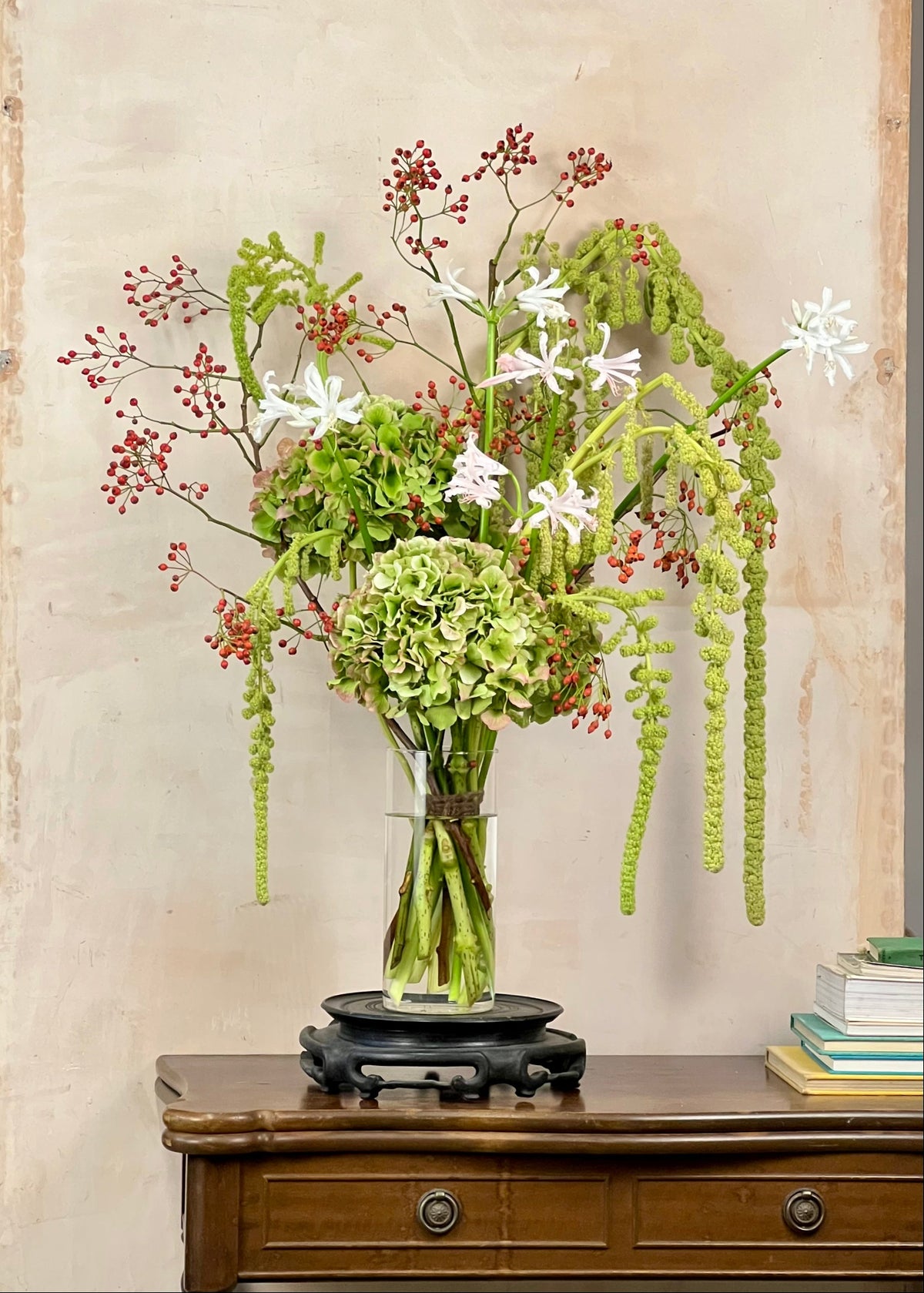 A floral arrangement in a clear glass vase with a black pedestal base, displayed on a wooden console table against a textured beige wall. The bouquet features green hydrangeas, trailing green stems, white star-shaped flowers, and clusters of small red berries.