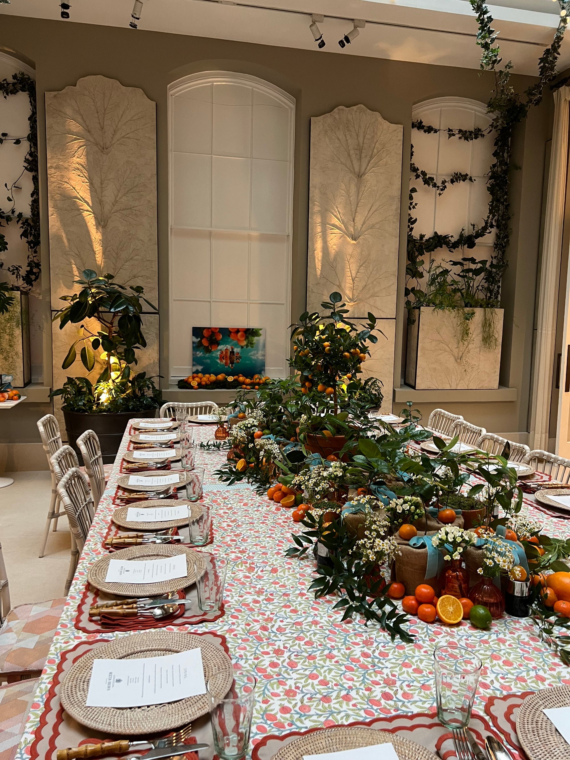 Decorated dining table with floral arrangements and a floral-patterned tablecloth. Orange themed plants and flowers with fruit on the table