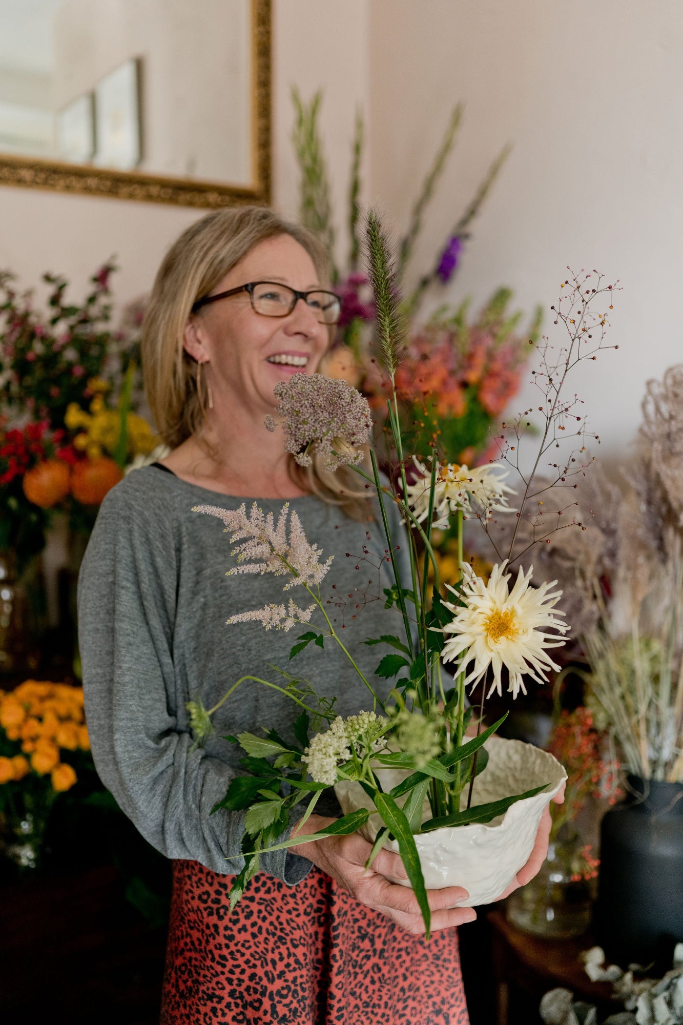 Woman holding a bowl of flowers in a room with floral arrangements