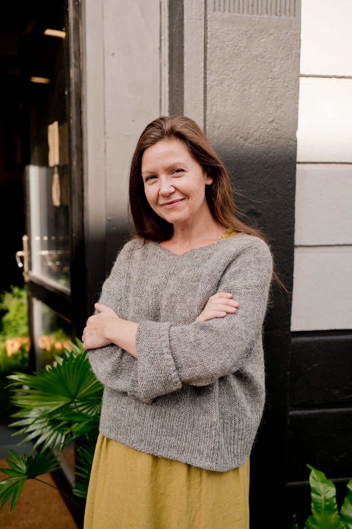 Woman wearing a gray sweater and yellow pants standing in front of a building with plants around.