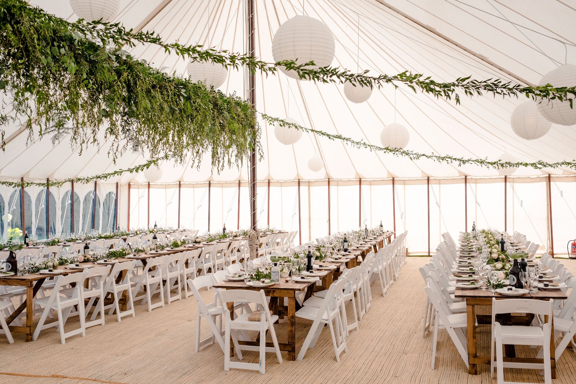 Decorated indoor event space with tables, chairs, and long hanging arrangement of foliage. slim lengths of foliage stretch from the central arrangement to the far corners of the marquee canopy. Greenery and flowers decorate the trestle tables