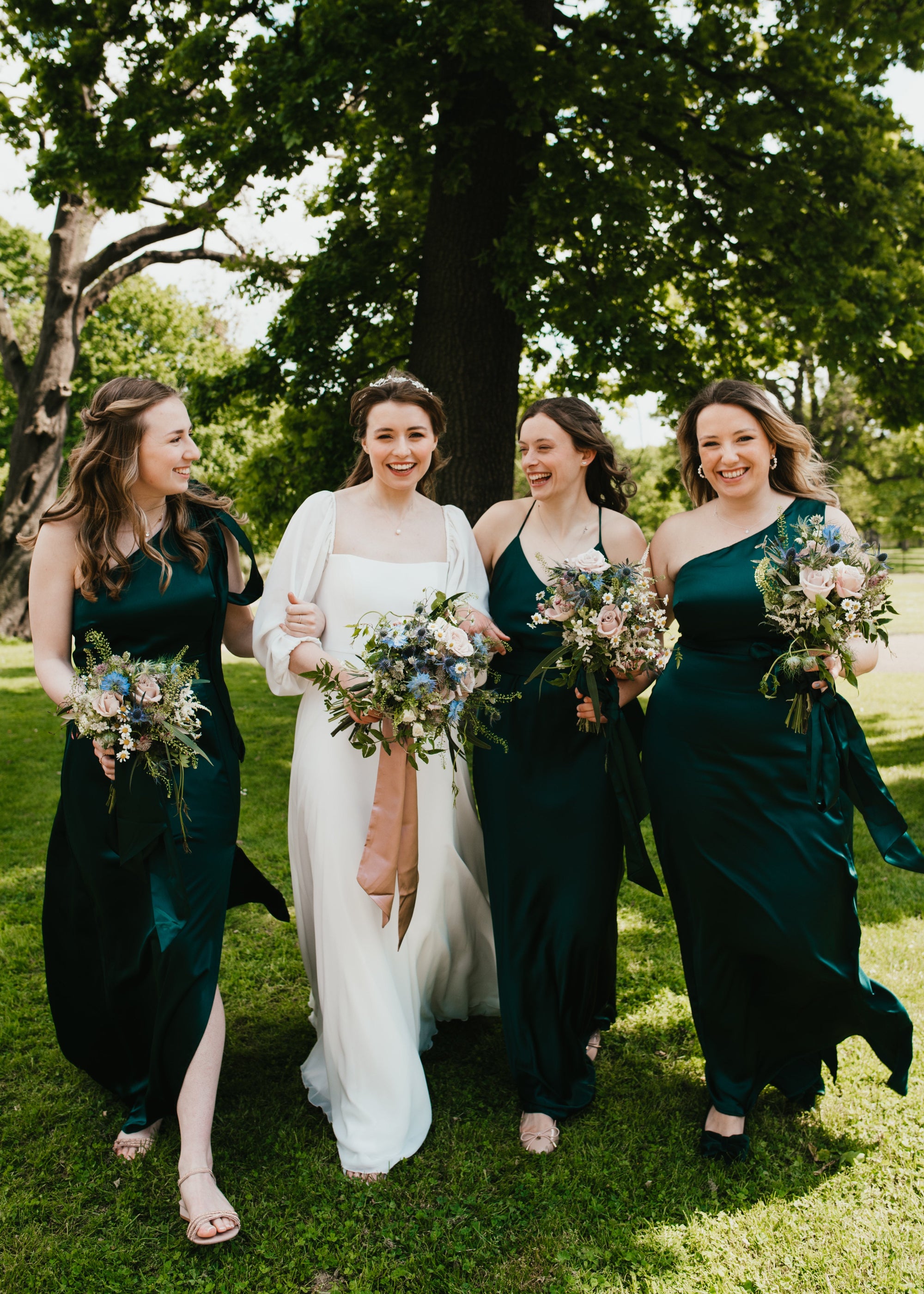 Bride with three bridesmaids in green dresses holding bouquets outdoors. Bouqiets are romantic in style with pale pink roses and thistles
