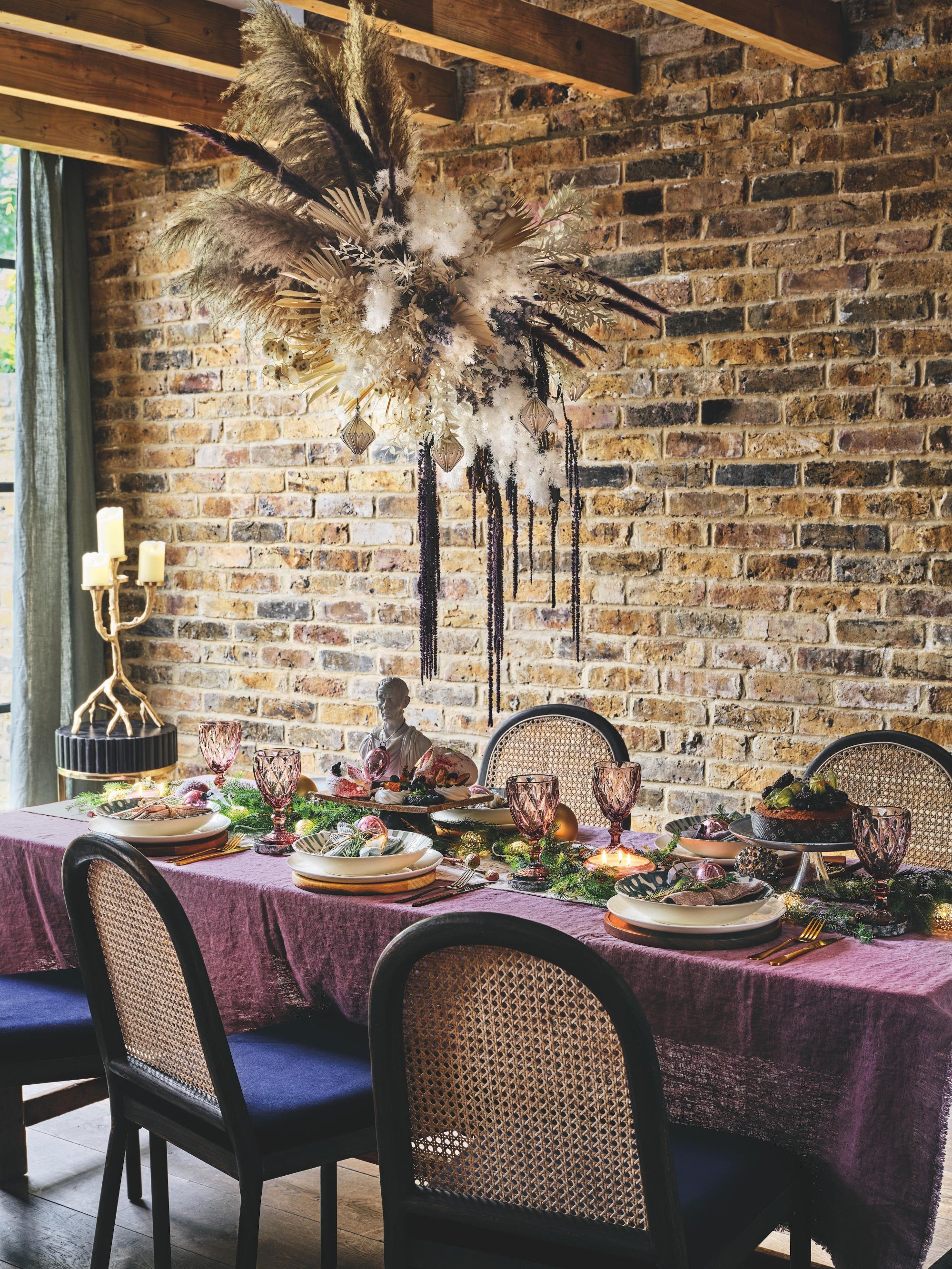 Table styling with plates, glasses, and candles against a brick wall. Dried floral handing arrangement with grasses and trailing dried flowers