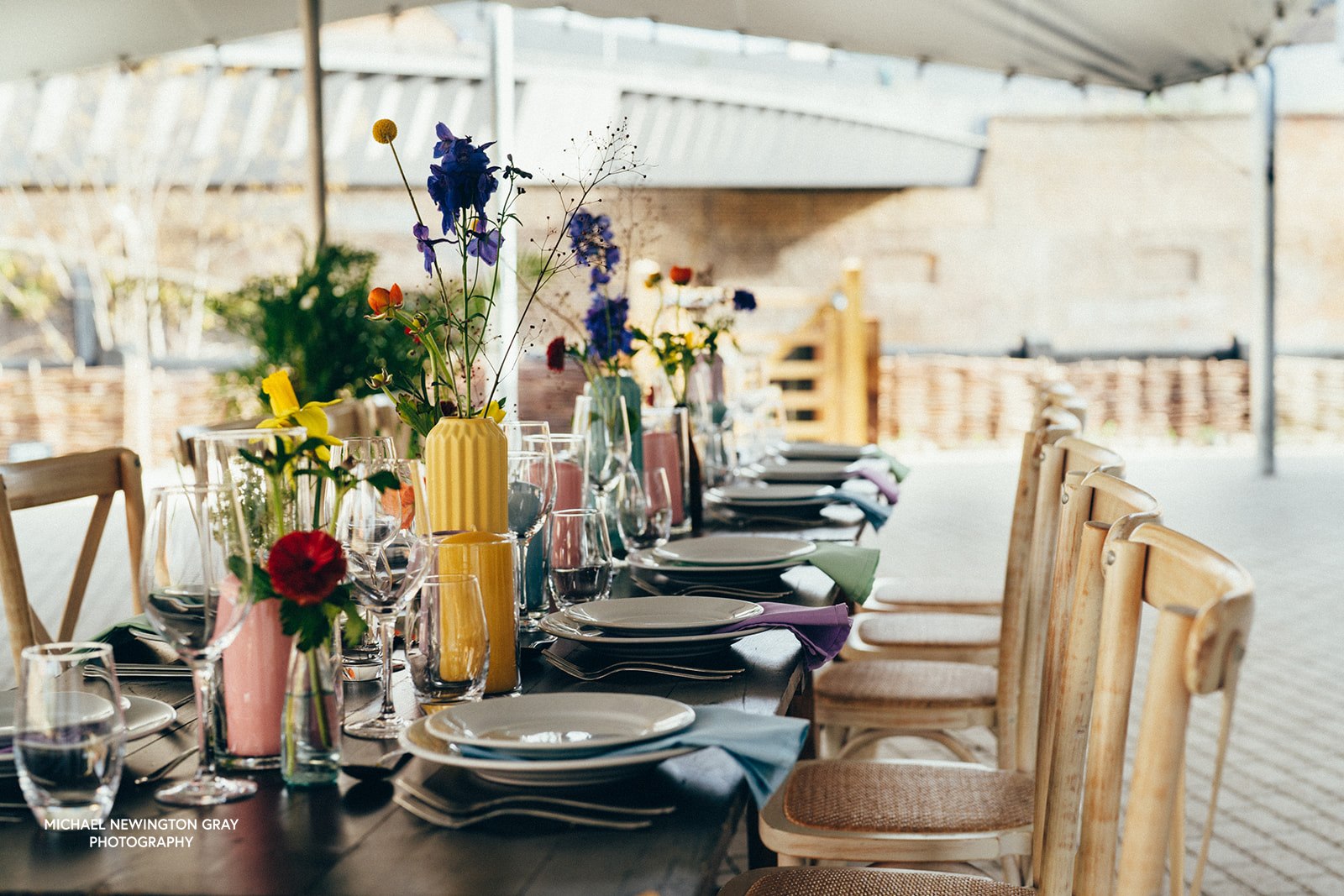 Dining table set with plates, glasses, and colourful floral arrangements in an outdoor setting under a canpy