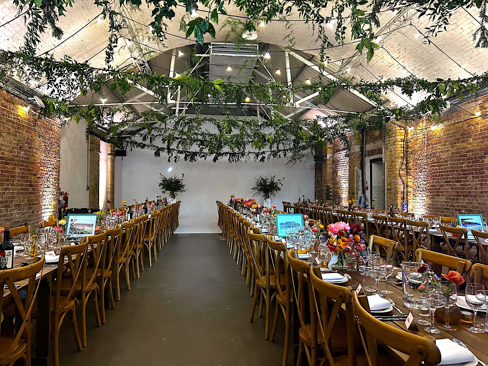 Long table set for a formal event with floral centre pieces in a room with brick walls and lots of fresh greenery hung from the ceiling. Pedestals of foliage in background