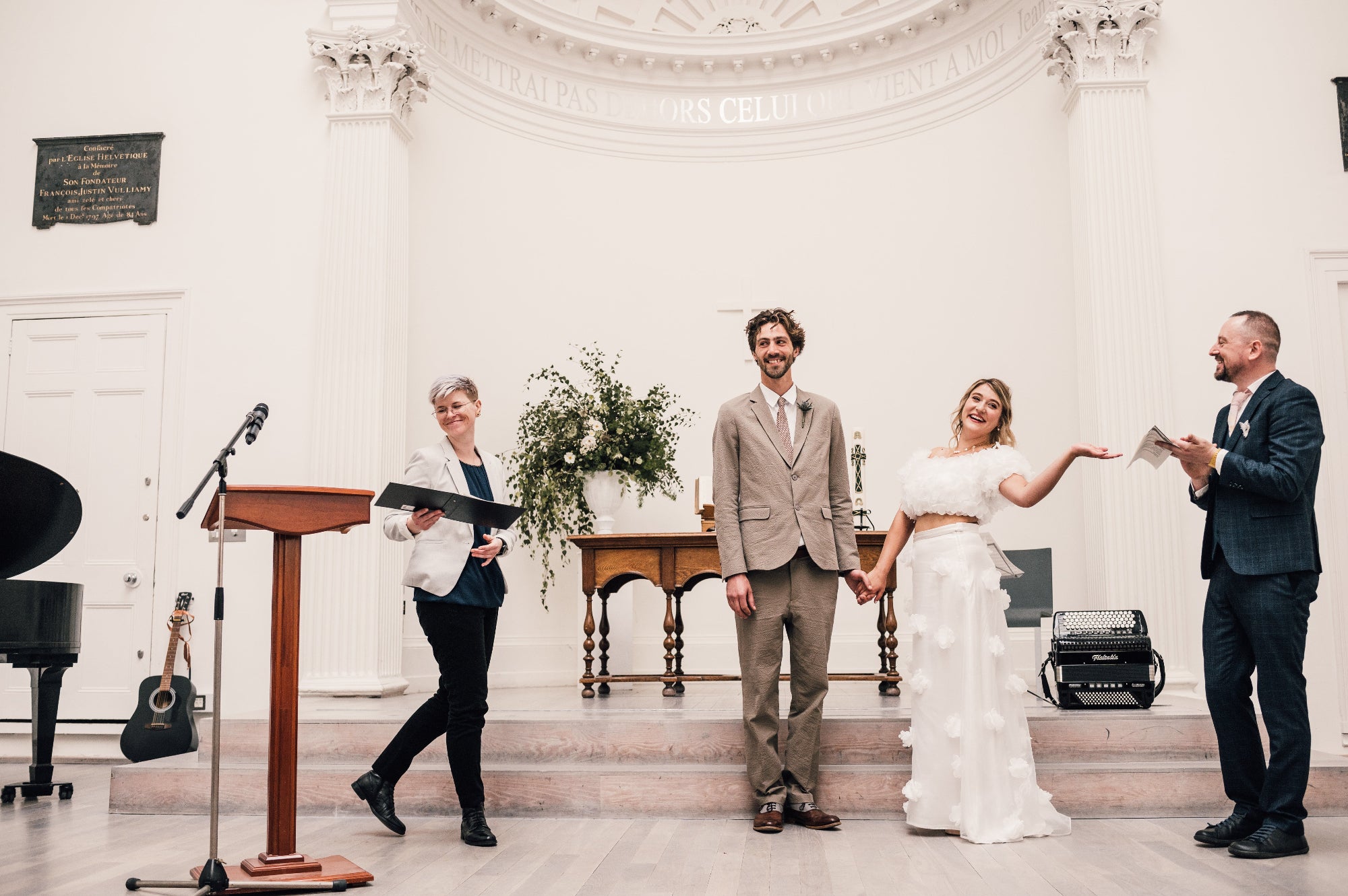 Wedding ceremony taking place in a high ceilinged formal setting with a couple and officiant. Large urn of foliage and white flowers behind them