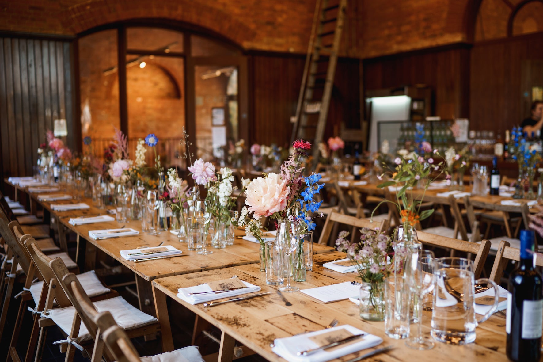 Long wooden tables set for a meal with floral centerpieces in a rustic indoor setting. Groups of bud vases scattered along the tables with pink peonies and blue delphinium
