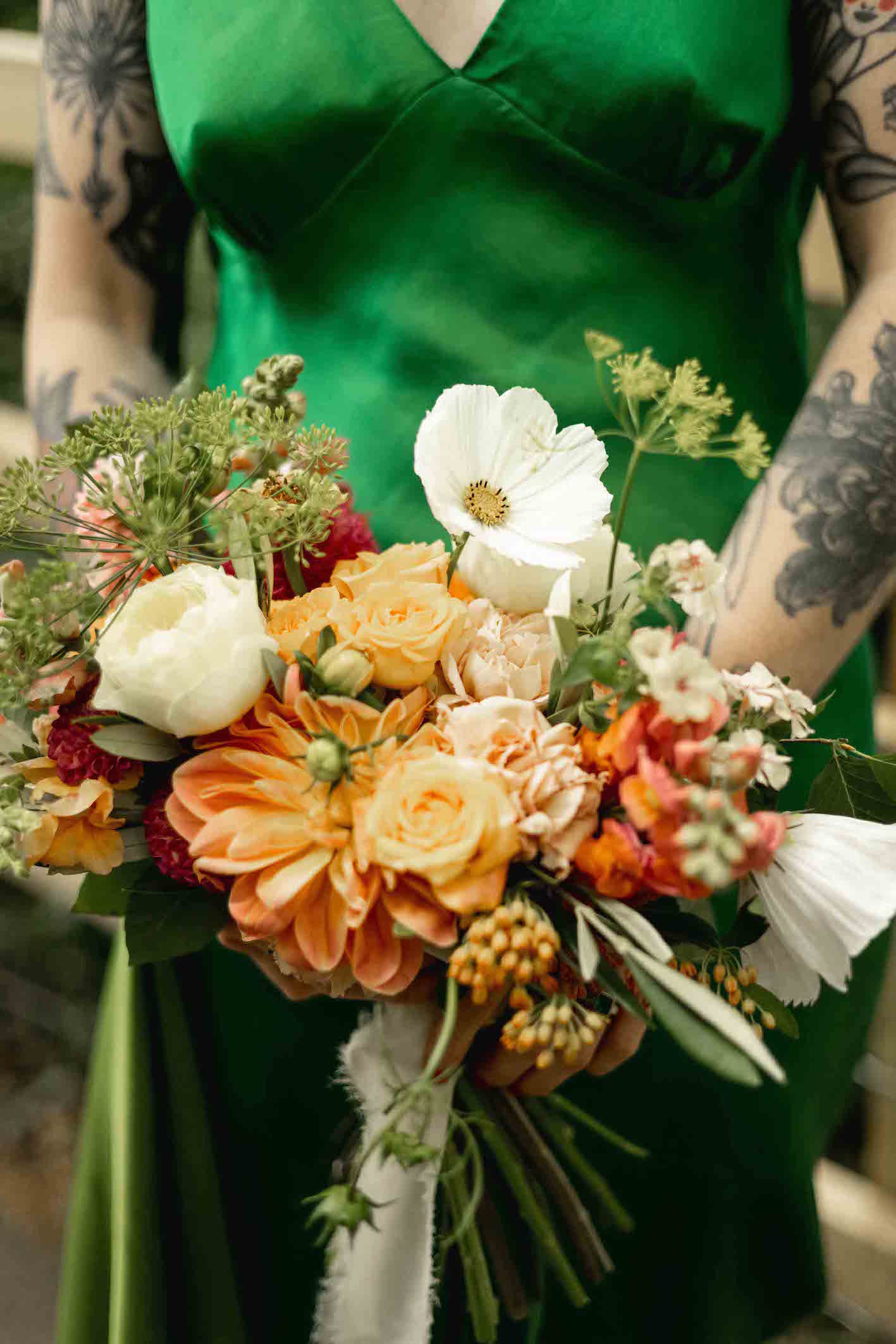 Bride wearing a silk green dress holding a colourful bouquet of flowers. Flowers are dahlia, cosmos, roses and dill in orange, white and yellow tones