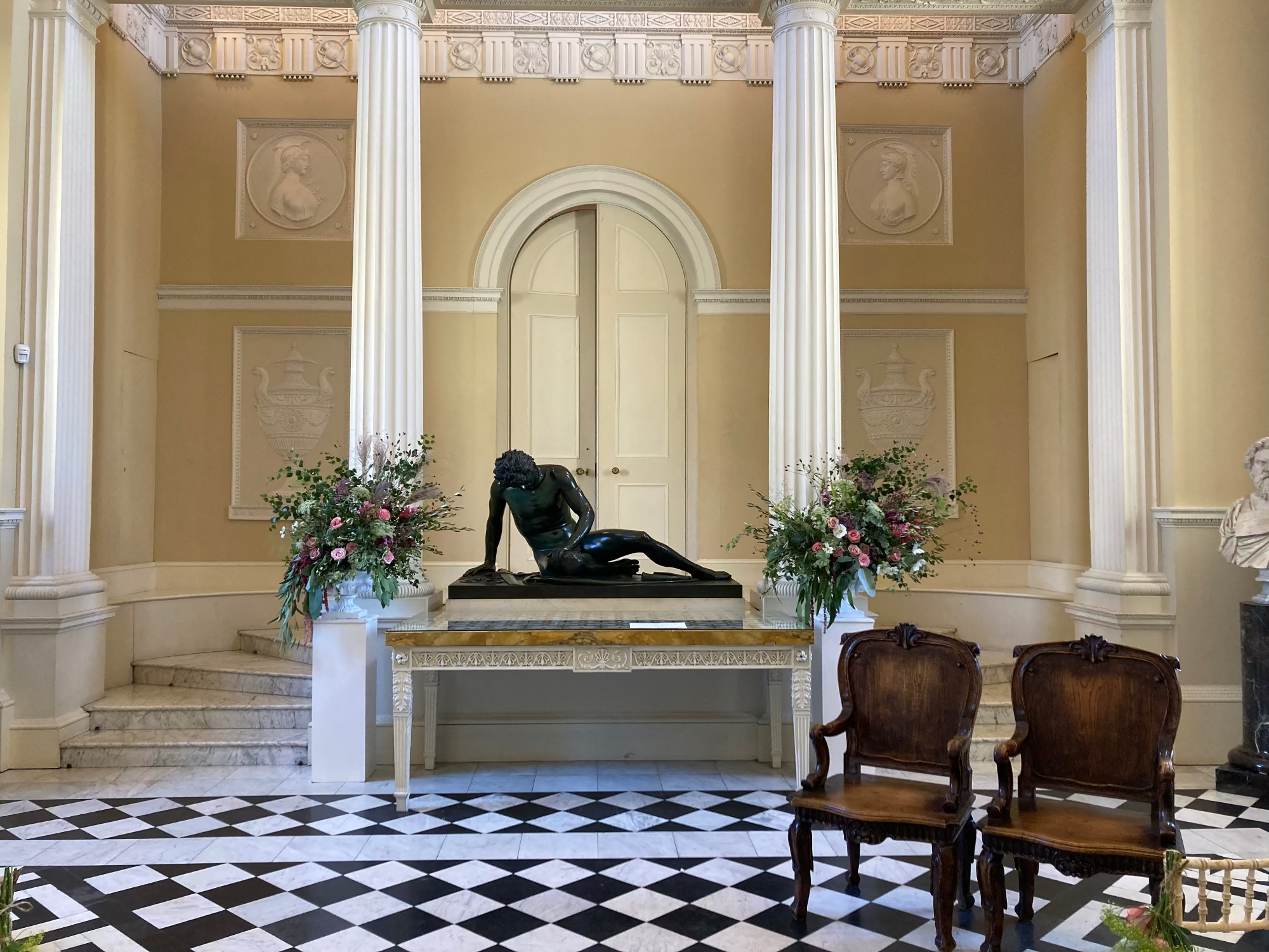 Decorative interior with classical statue, floral arrangements, and marble columns. Large floral arrangement in white urns with pink, green and burgundy flowers and foliage