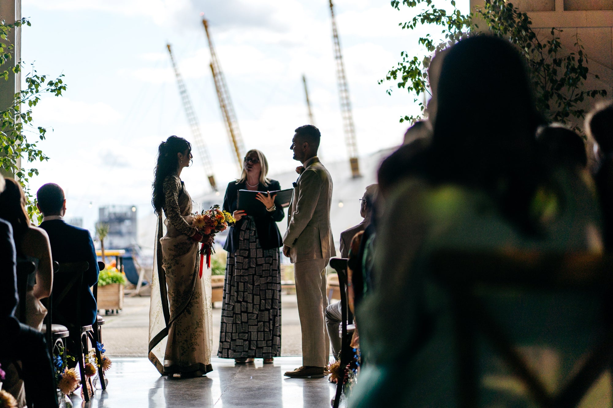 Wedding ceremony taking place with a couple with 02 in the background. Trees frame the couple and the bride holds a colourful bouquet
