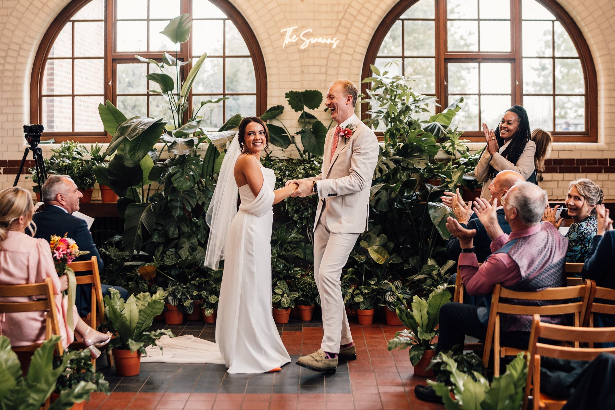 Wedding couple holding hands in front of guests in an industrial room with large windows. A large backdrop of house plants creates a focal point for the ceremony