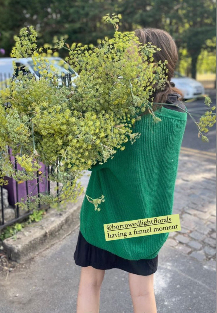 Florist wearing a green jumper with floral design and text, holding flowers outdoors. Large bunch of Dill over shoulder