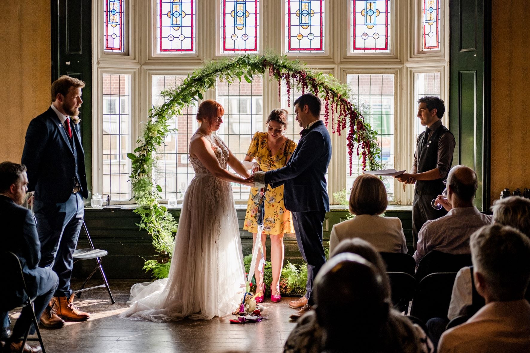 Wedding ceremony taking place in a room with stained glass windows. Couple are holding hands in front of a floral moon gate decorated with delicate green foliage and deep red trailing flowers
