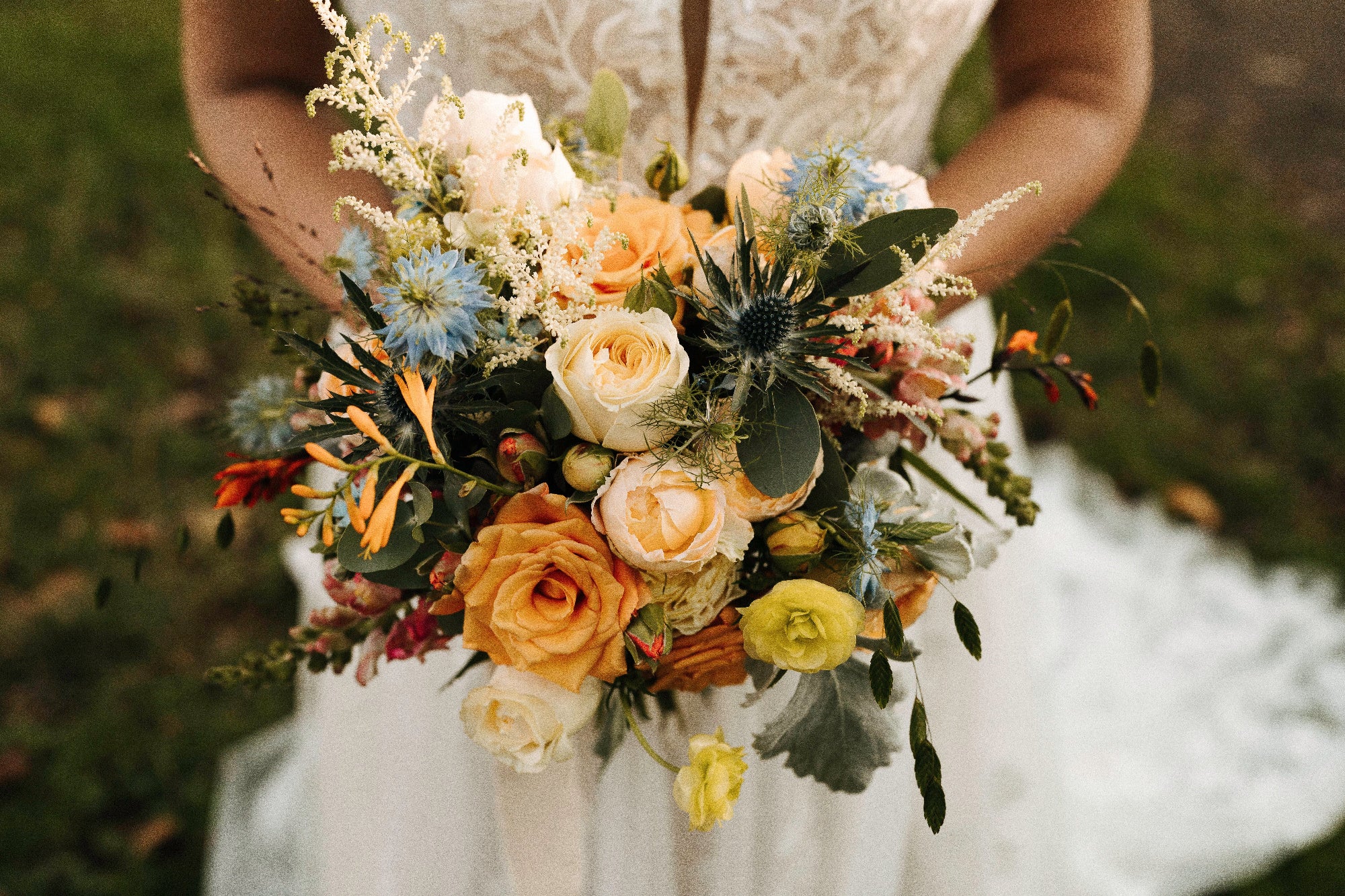 Bouquet of flowers held by a bride wearing a lace dress. Pastel flowers in the bouquet include cream roses, thistle, yellow butterfly ranunculus, nigella and Crocosmia