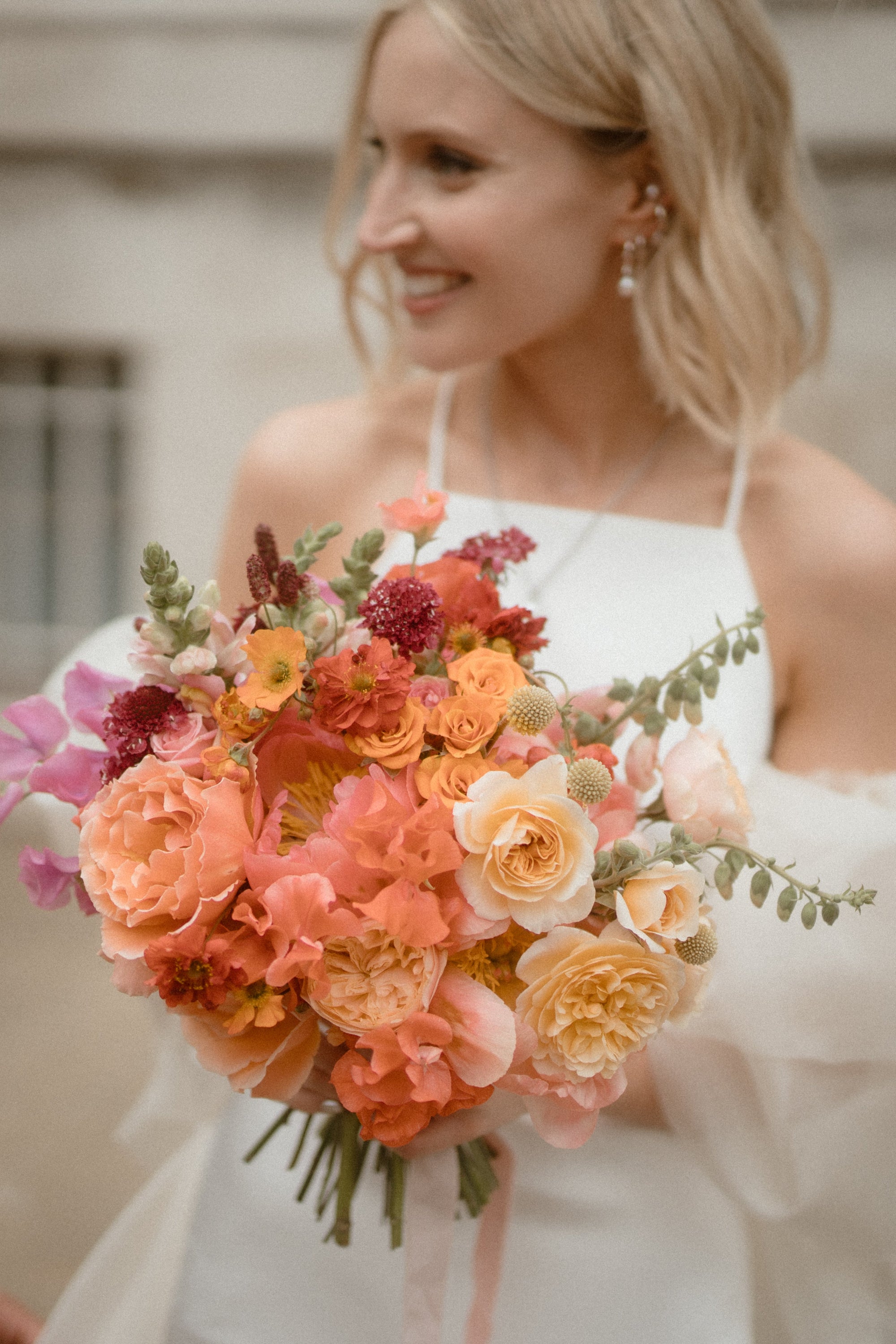 Bride holding a colourful peach and pink bouquet with a blurred background. flowers include roses, sweet peas, and scabious