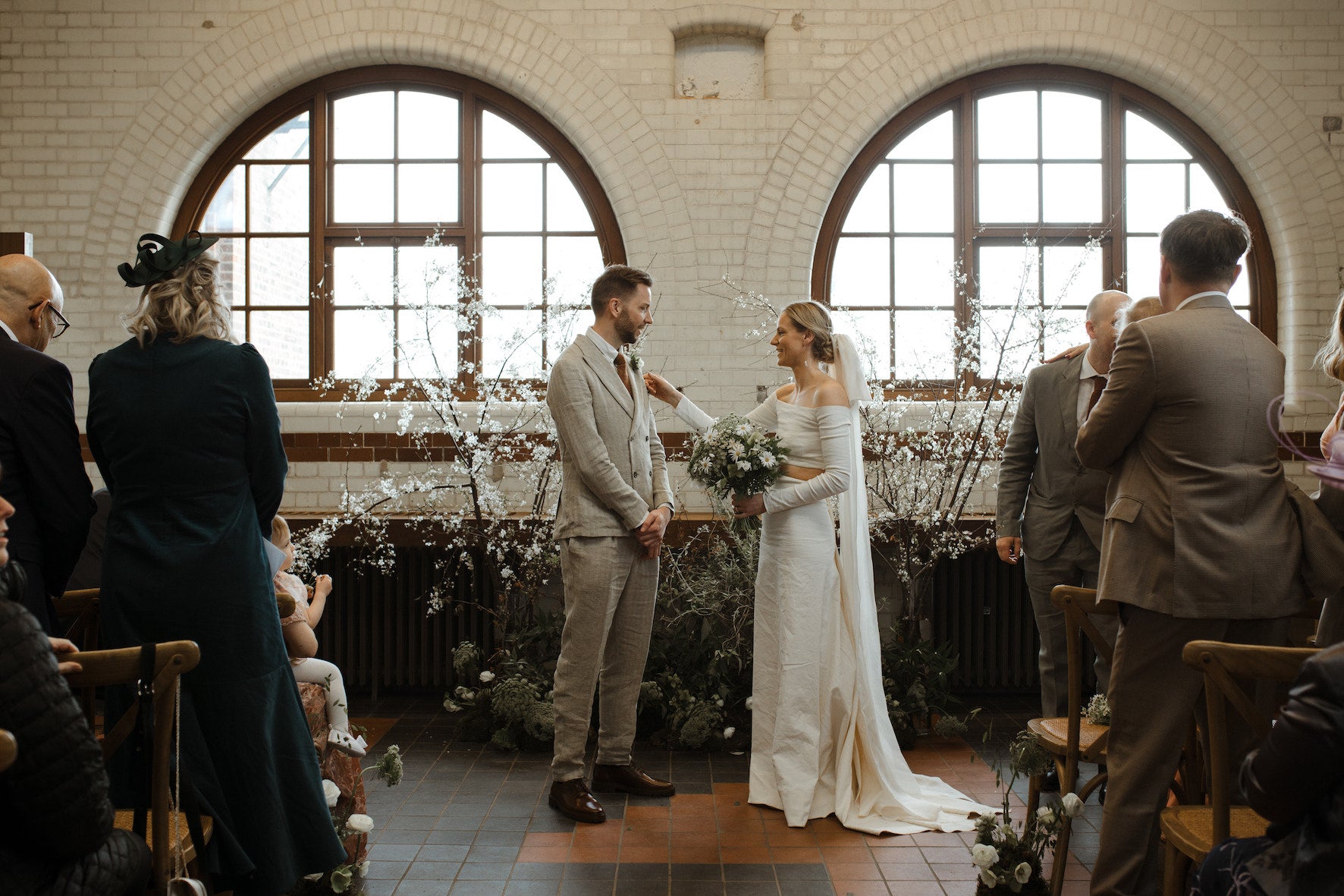 Wedding ceremony taking place in an industrial room with large windows and brick walls. Floral backdrop of white blossom with silver green foliage at the base. Bride holds a bouquet of fresh daisies and cow parsley