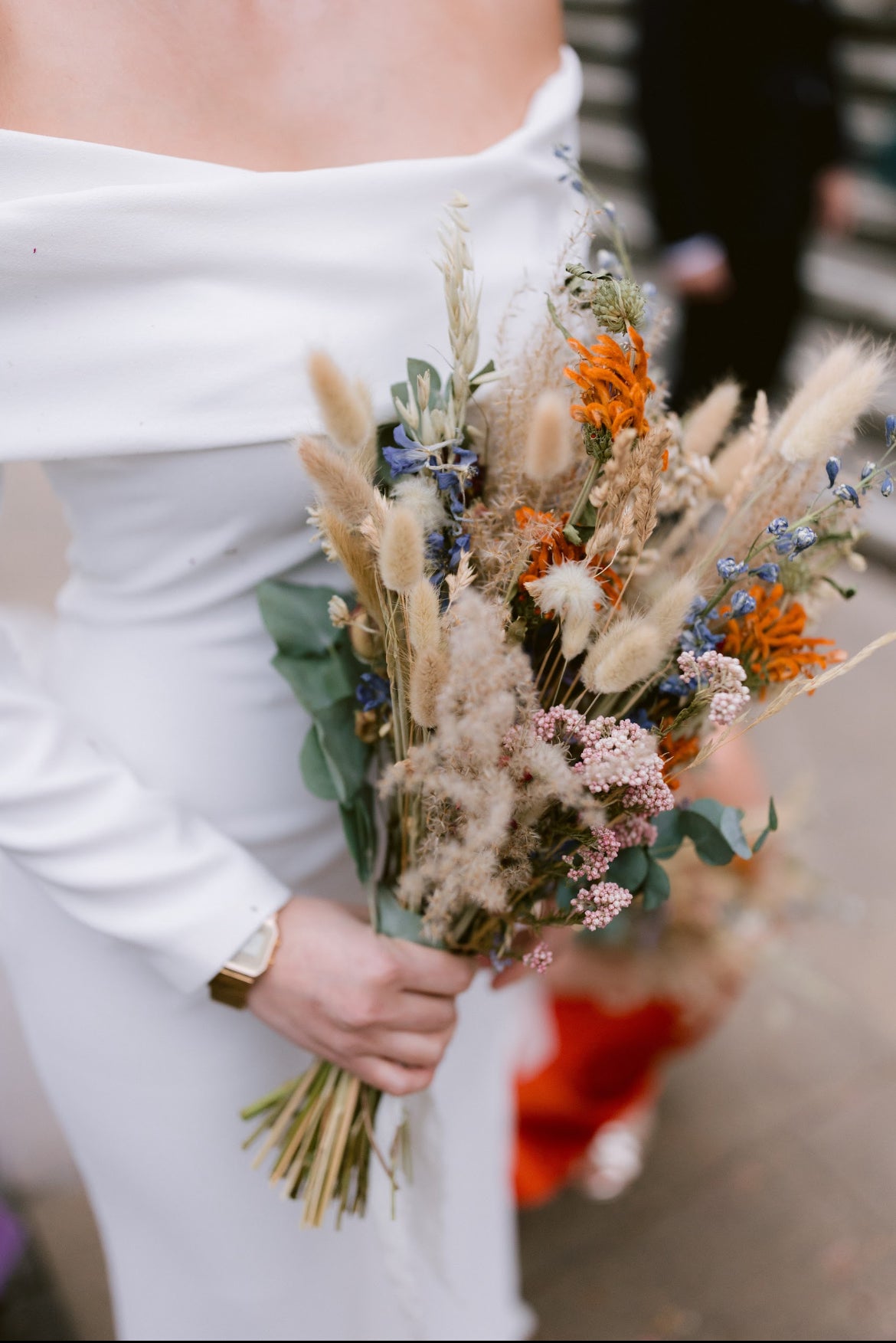 Bouquet of dried flowers held by a bride in a white dress. Dried pink, orange, blue and neutral coloured flowers and grasses