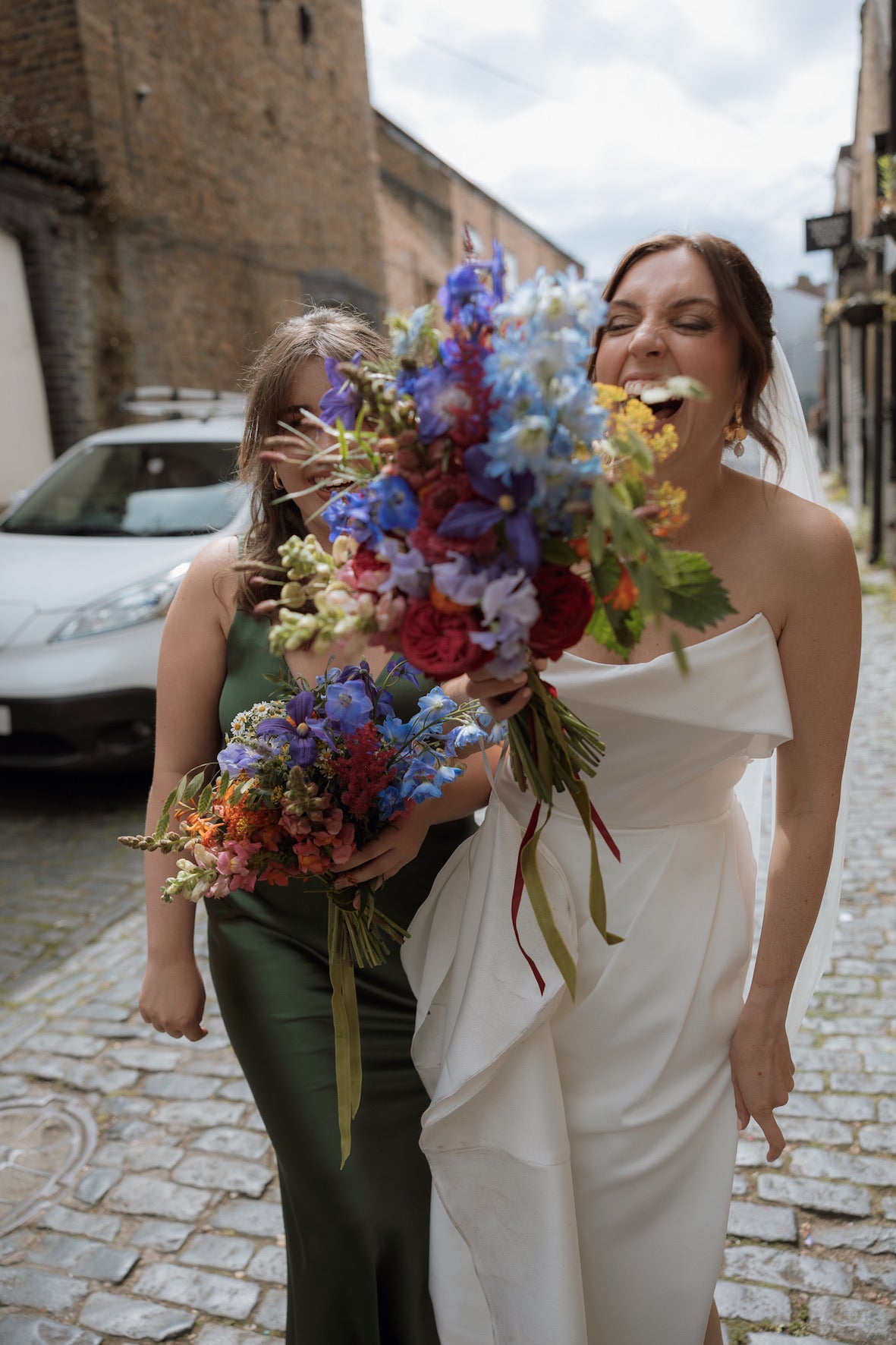 Bride and bridesmaid holding colourful flower bouquets on a cobblestone street in east London. Bride looks ecstatic whilst looking at her flowers. Bouquet is bright summer colours and includes delphinium, scabious and antirhinum