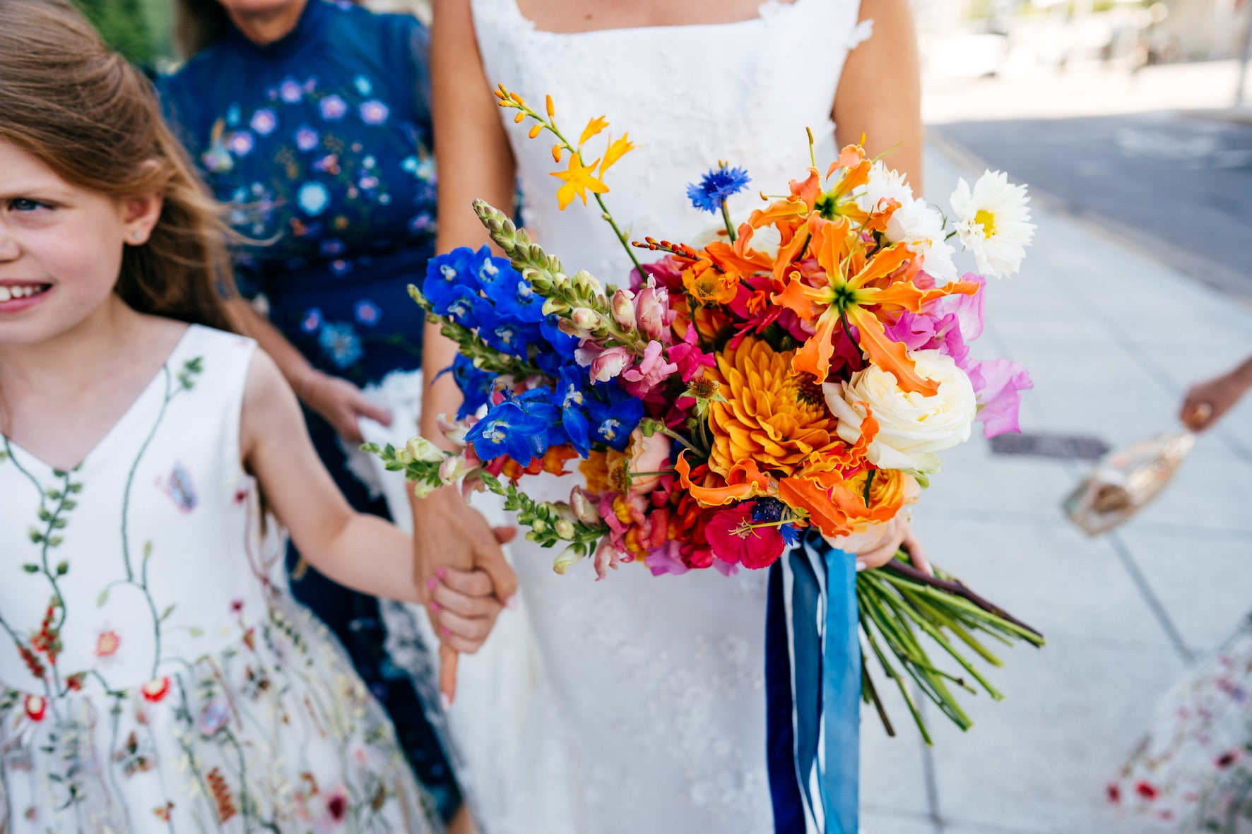 Bride holding a colourful bouquet with a young girl in a floral dress. Bright blue delphinium, orange dahlia, Gloriosa lily and Crocosmia, Antirrhinum and white cosmos