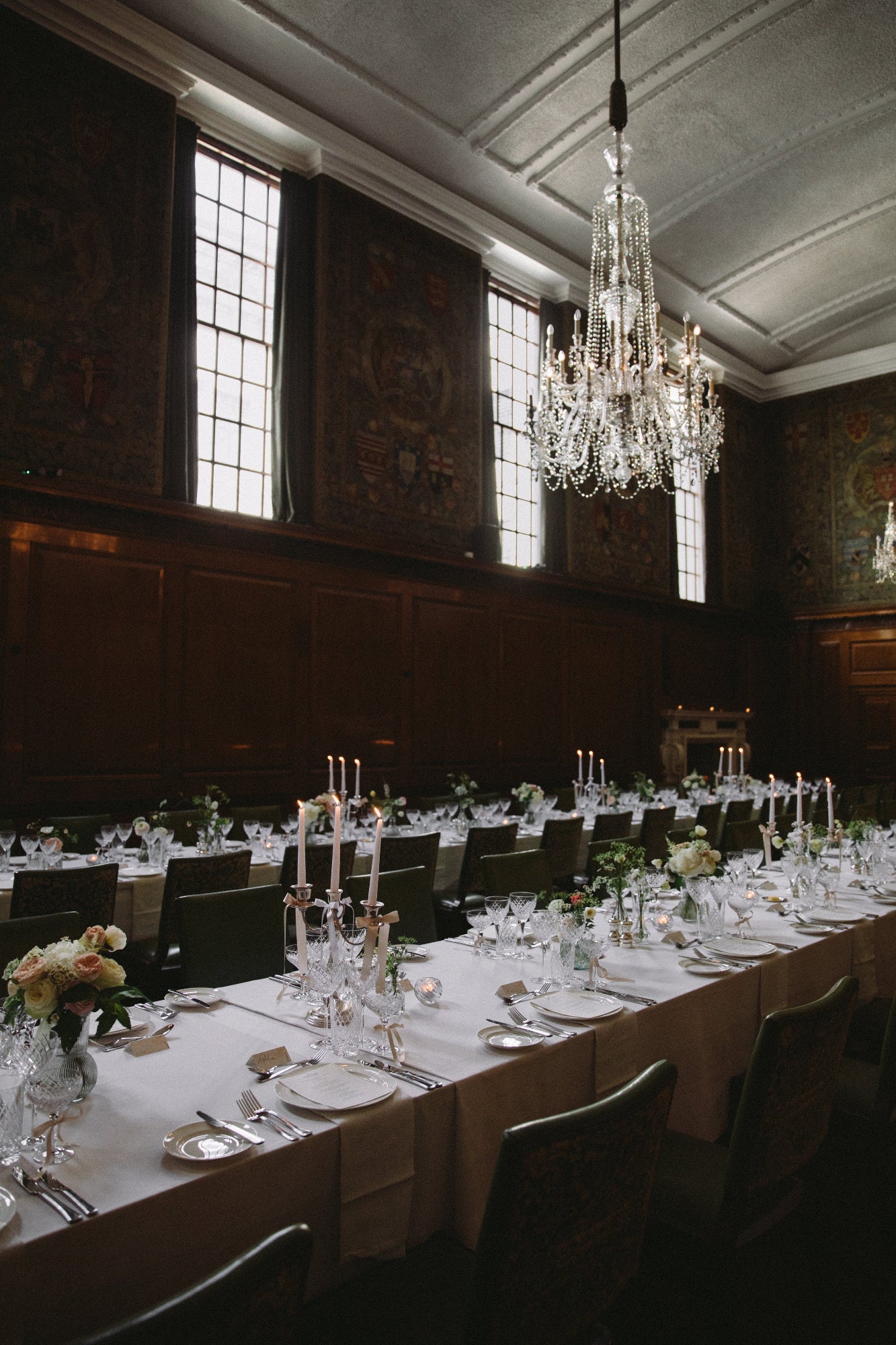 Grand wooden panelled dining room with long table set for a formal meal, chandelier hanging from ceiling. Bud vases of delicate and romantic flowers decorate the table with tall tapered candles and silver candlestick holders