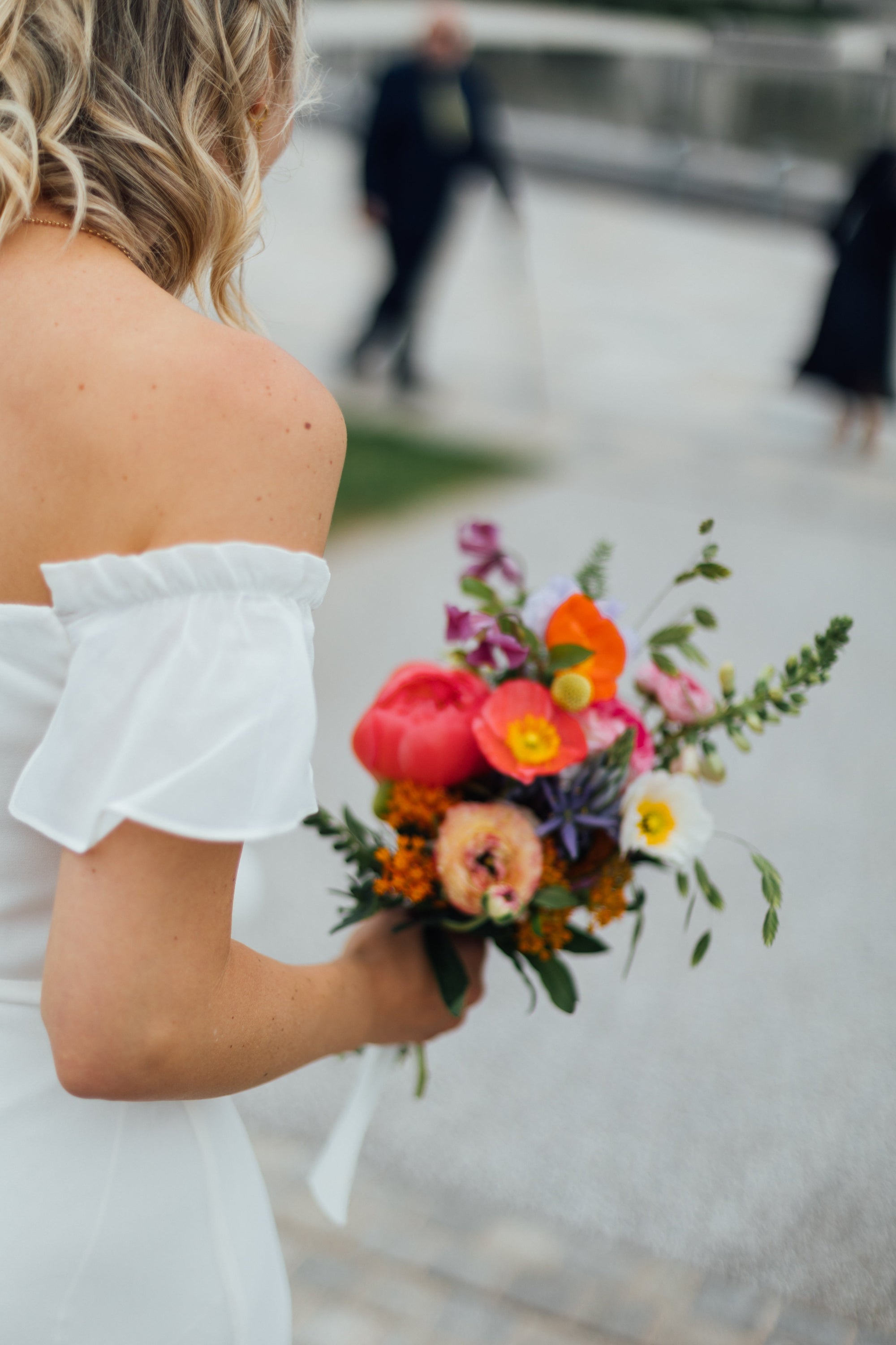 Bride holding a colourful bouquet of coral poppies and peonies with blurred background. Bouquet has thistles and grasses with some berries