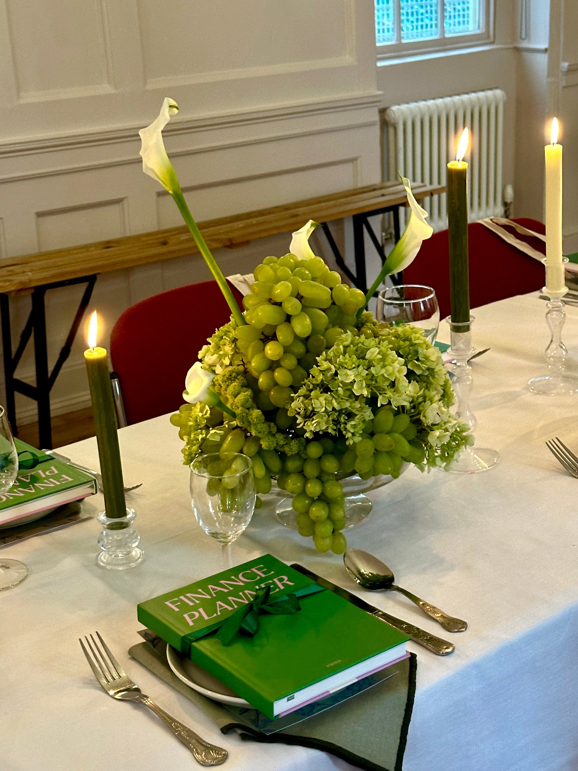 Table setting with green centerpiece of grapes, hydrangea and calla lillies, Table decoration include books, and candles in a formal dining room.