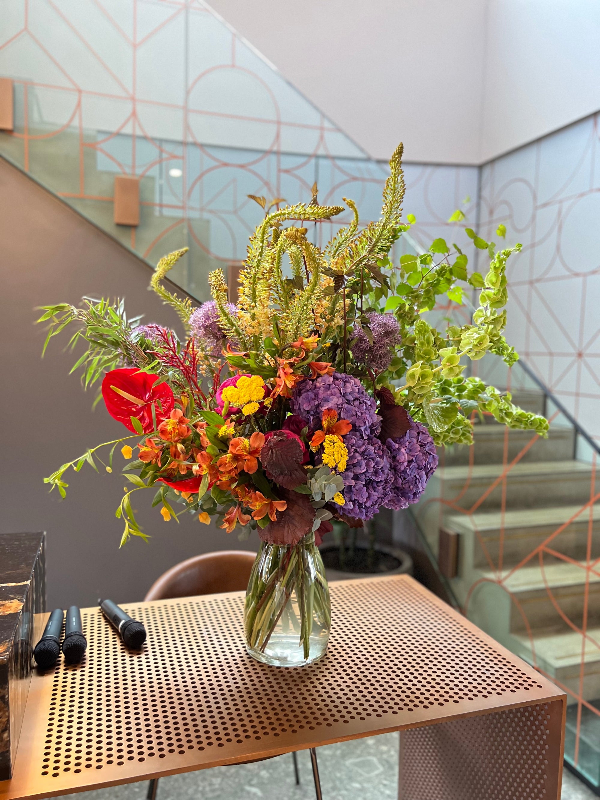 Colorful floral arrangement in a clear vase on a wooden table with a modern interior background. Includes hydrangea and anthurium