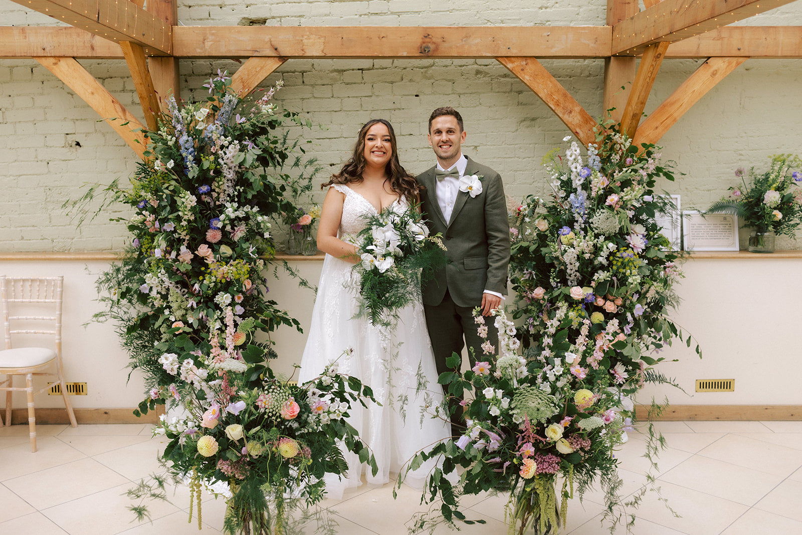 Couple standing between large floral arrangements in a rustic indoor setting. Floral column of pastel garden flowers including roses, delphinium, dahlia, sedum and amaranthus. Bridal bouquet of foliage and white orchids