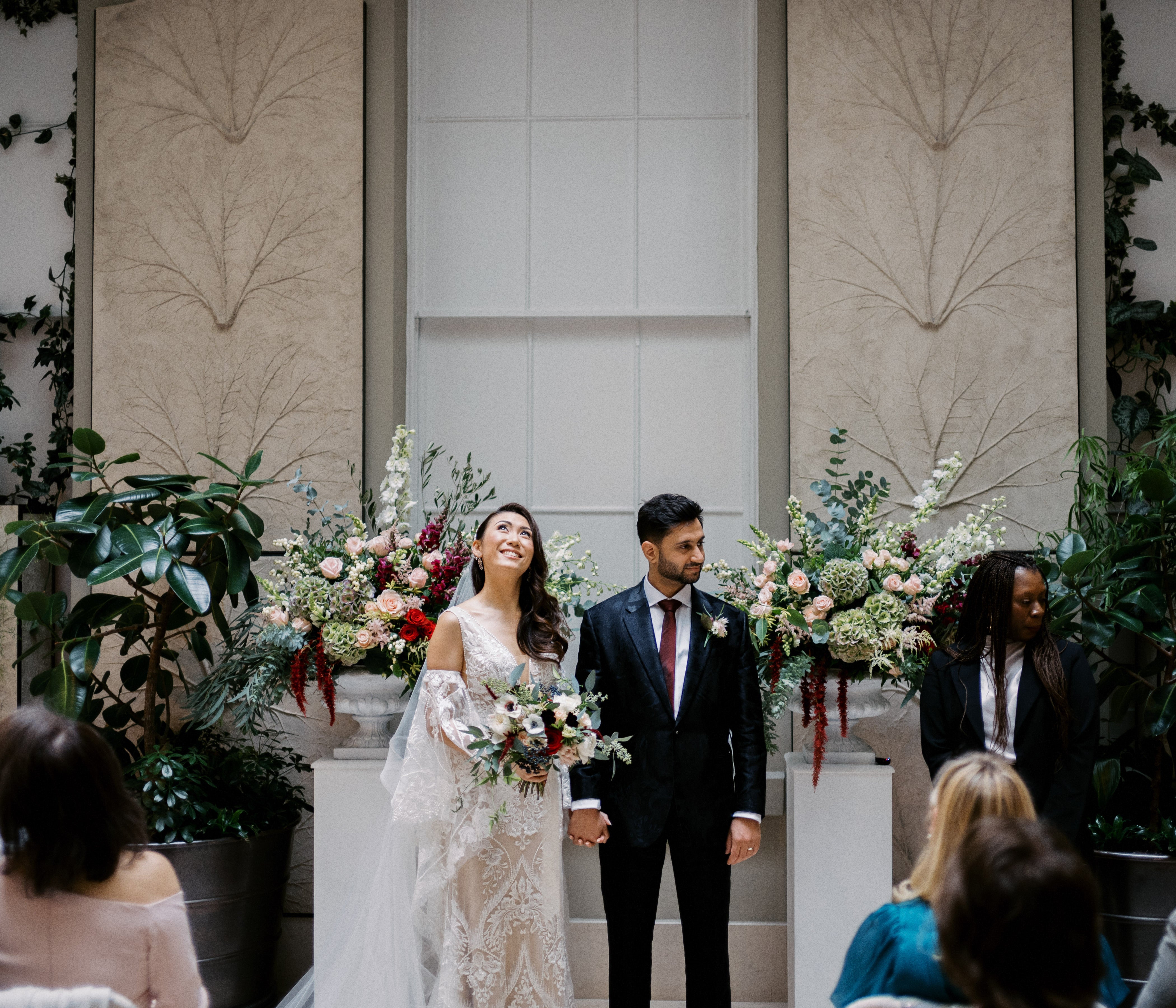 Wedding couple standing in front of large flora pedestals with guests in the foreground. Bride holds bouquet of white and burgundy anemone. Flowers are arekred, white, green and blush pink.